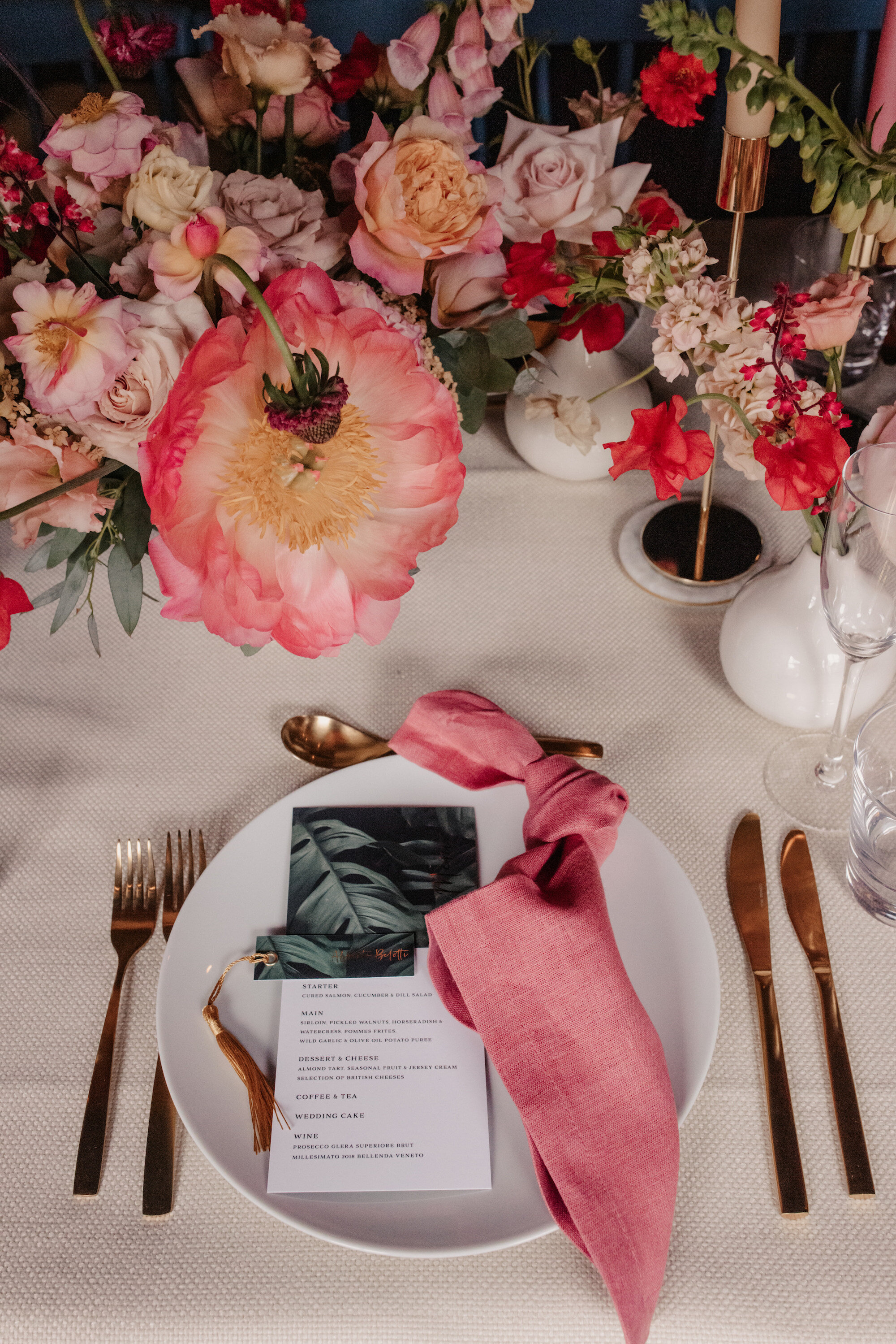 A colourful place setting of a London wedding with pink linen napkins, gold cutlery, and red and blush centrepieces