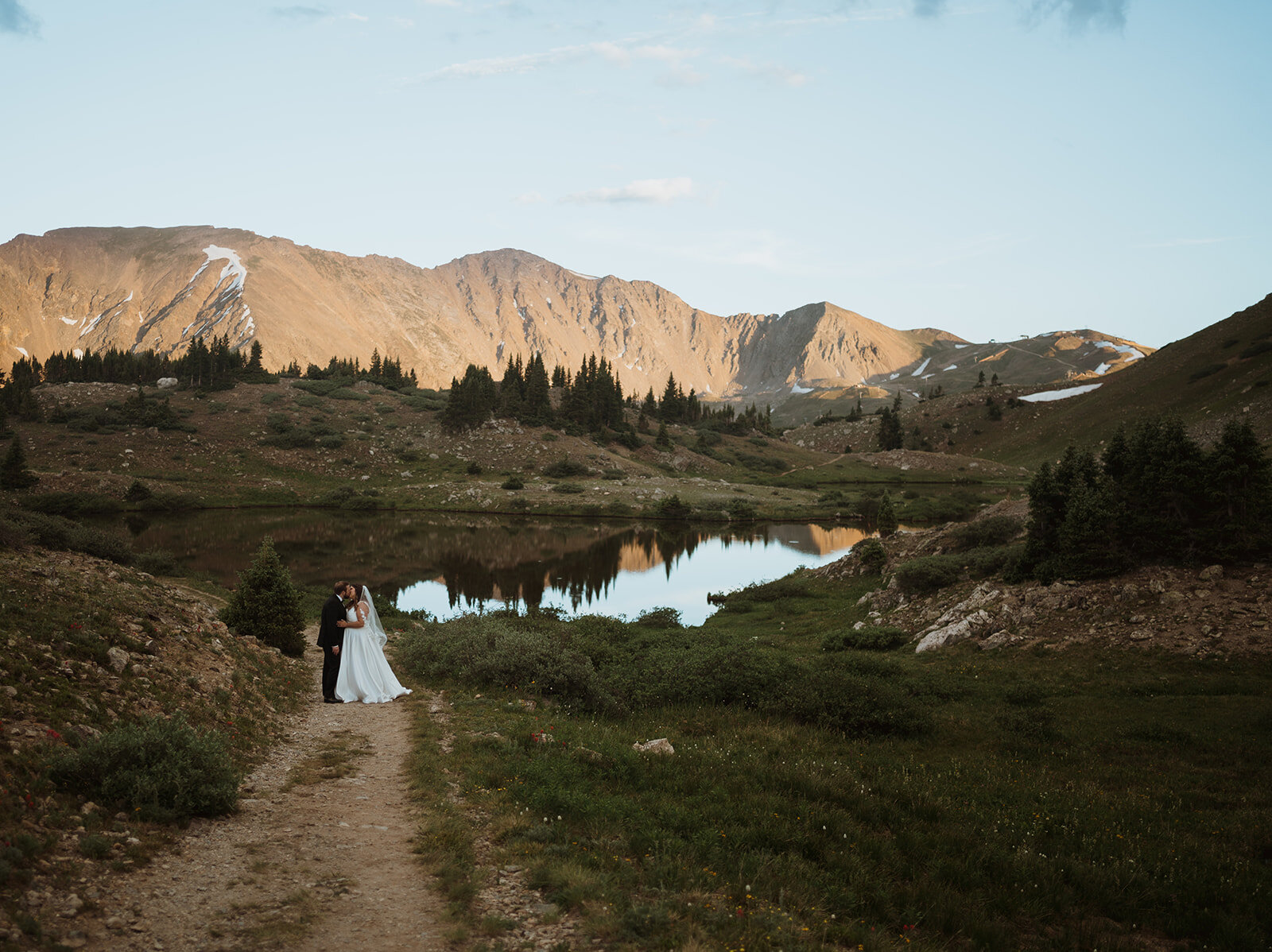 bride and groom are embracing in front of a huge mountain range as the sun sets. there is an alpine lake in the background and the shadows of the mountains are getting cast on it. there is green grass everywhere and rocks.