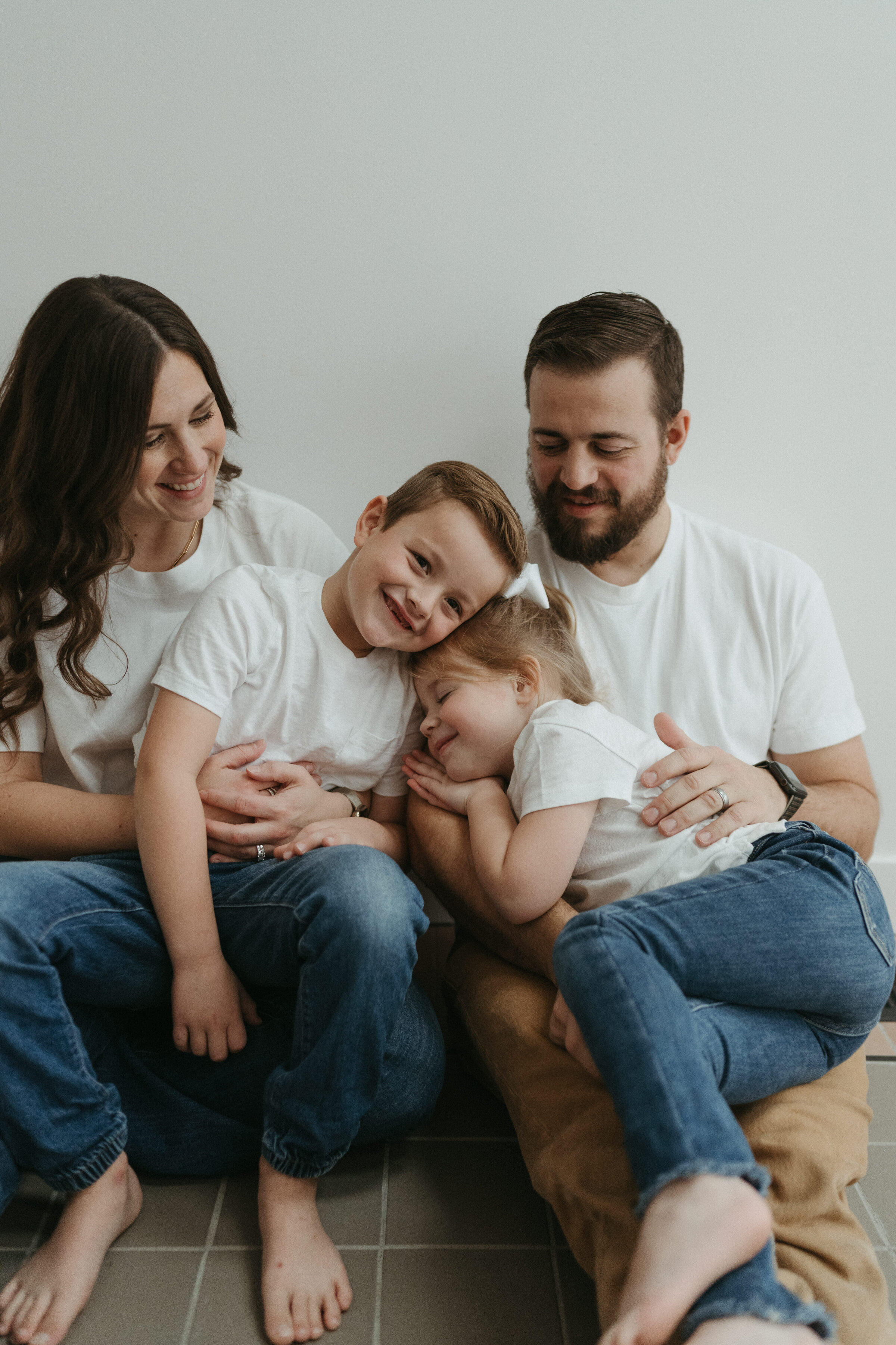 A mom, dad, young boy and toddler girl all in matching white shirts, snuggle in a group on a stone floor in front of a white backdrop.