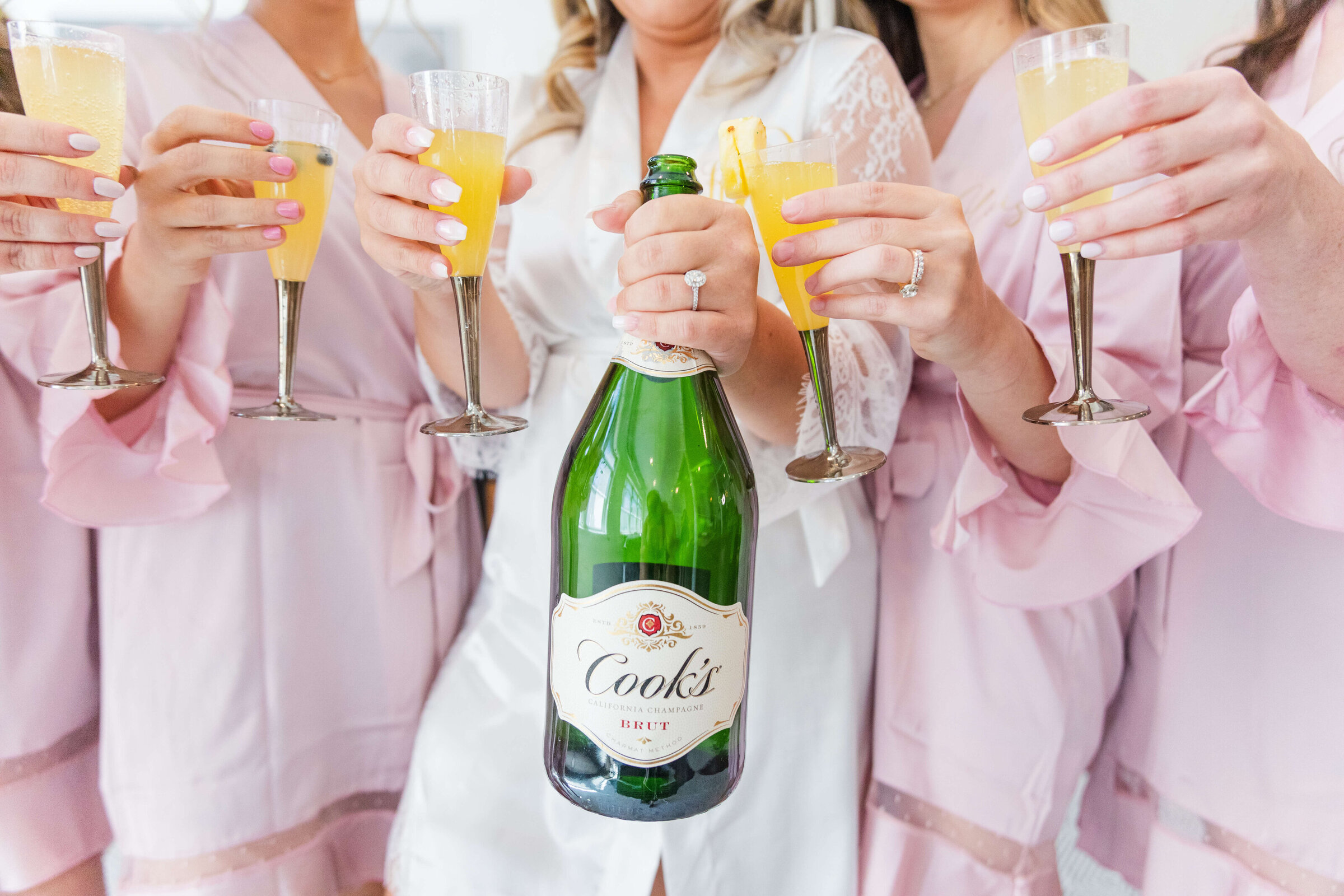 A bride is in a white robe and holds up a bottle of champagne while bridesmaids in pink robes hold up their mimosas