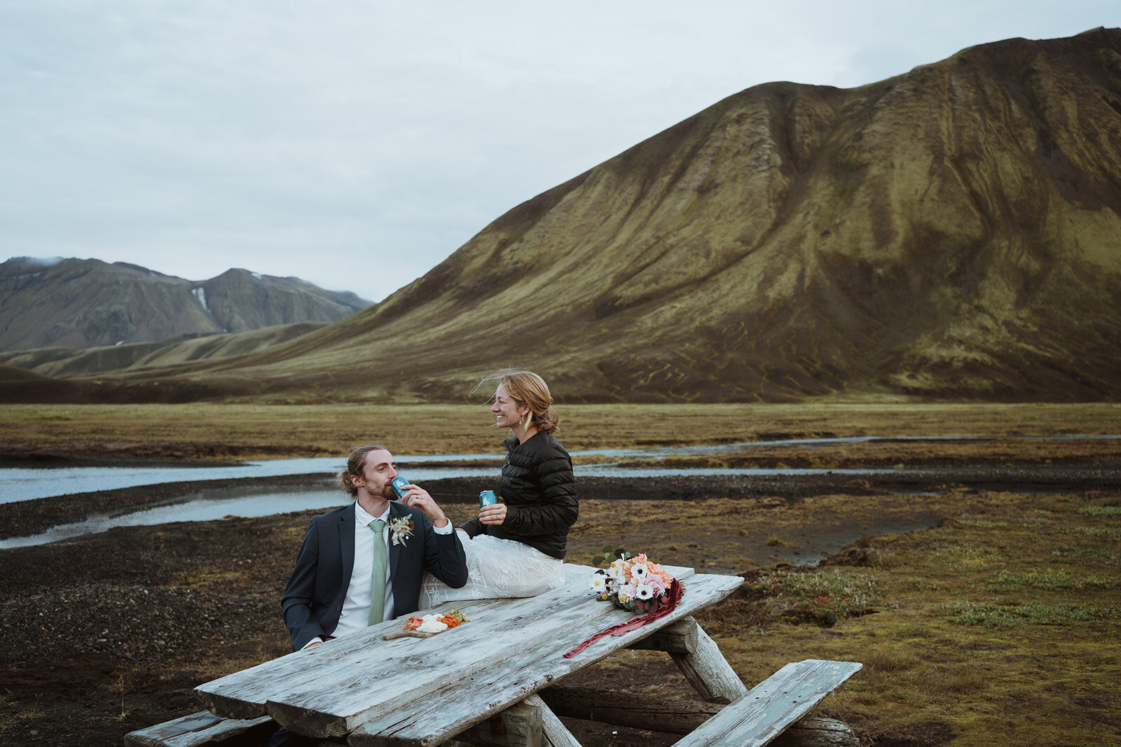 the bride and groom are having a picnic after their iceland elopement. they have craft beers, and have a charcuterie board. the bride is sitting on the picnic bench, and the groom is sitting next to her. she is looking out at the scenery and the groom is sipping his drink.