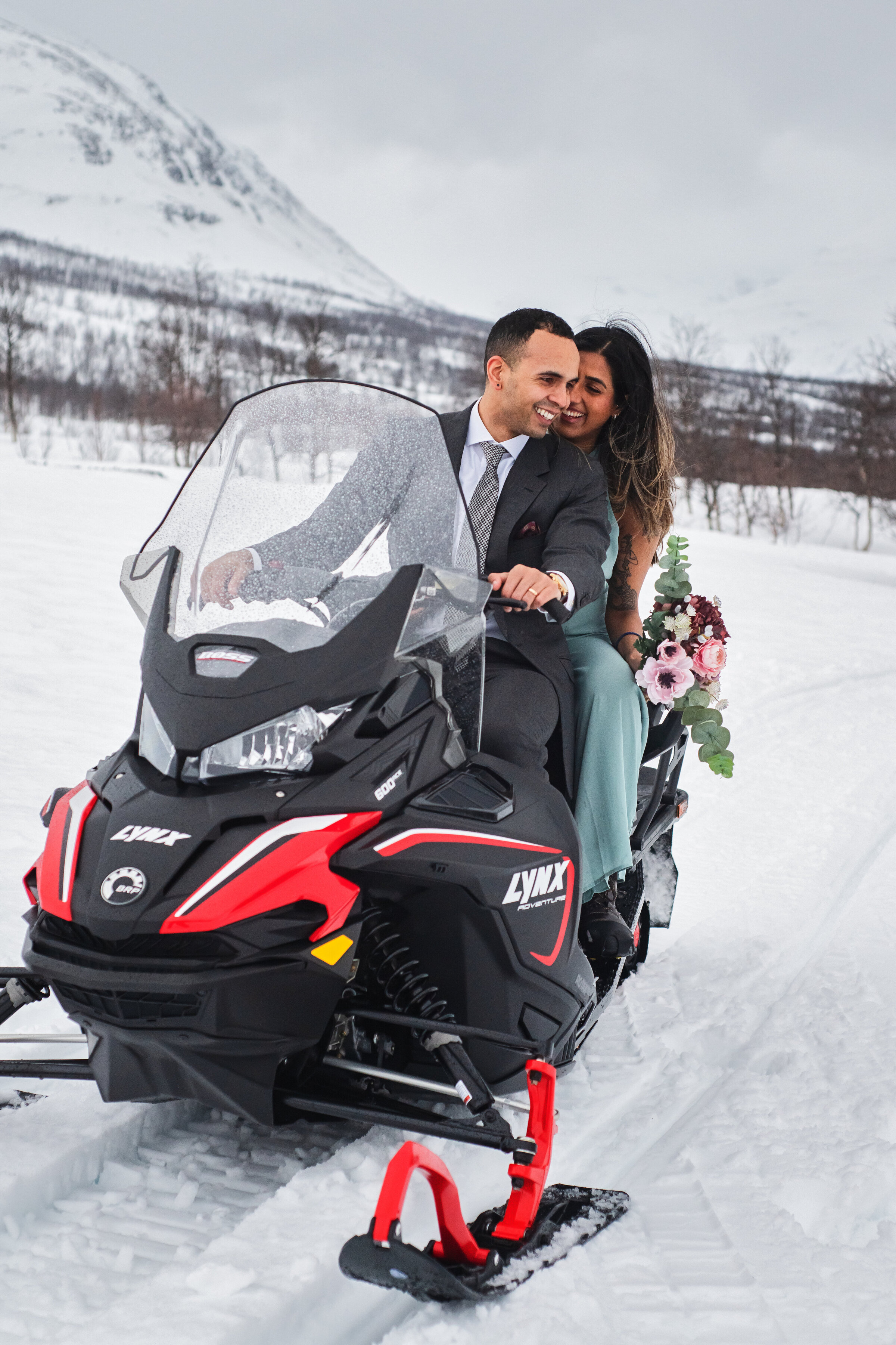 A newly eloped Indian couple sharing a private snowmobile trip, smiling at each other on a snowmobile, with the groom in a suit and the bride in a teal dress, set against a snowy Norwegian mountain landscape. The groom is driving and the bride riding passenger behind carrying a bouquet of flowers.