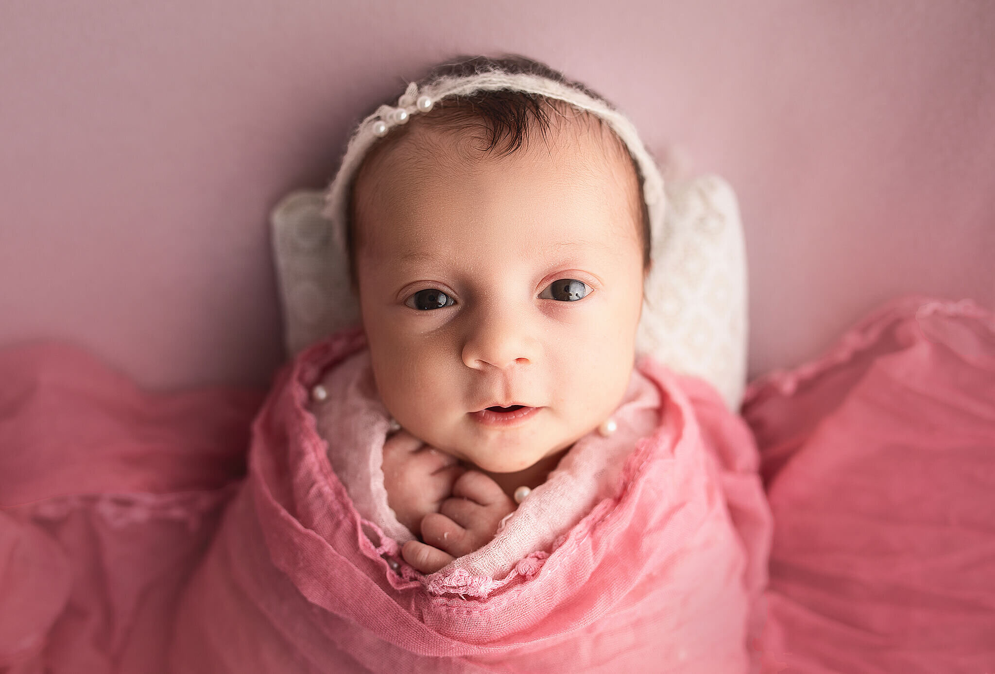 Newborn girl wrapped in pink wrap, laying head on pillow and looking straight at camera