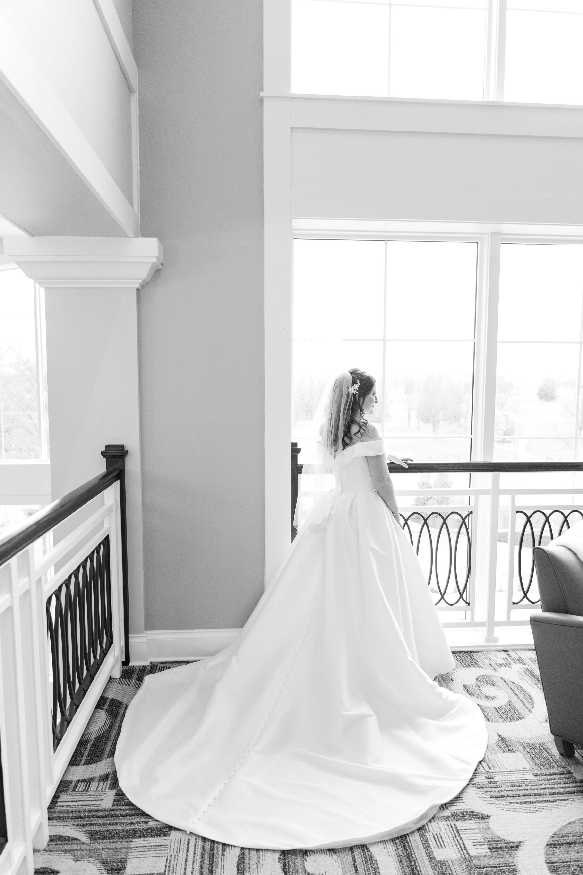 A black and white photo of a bride looking out a window. It's a wide shot, so her whole dress and train are in view.