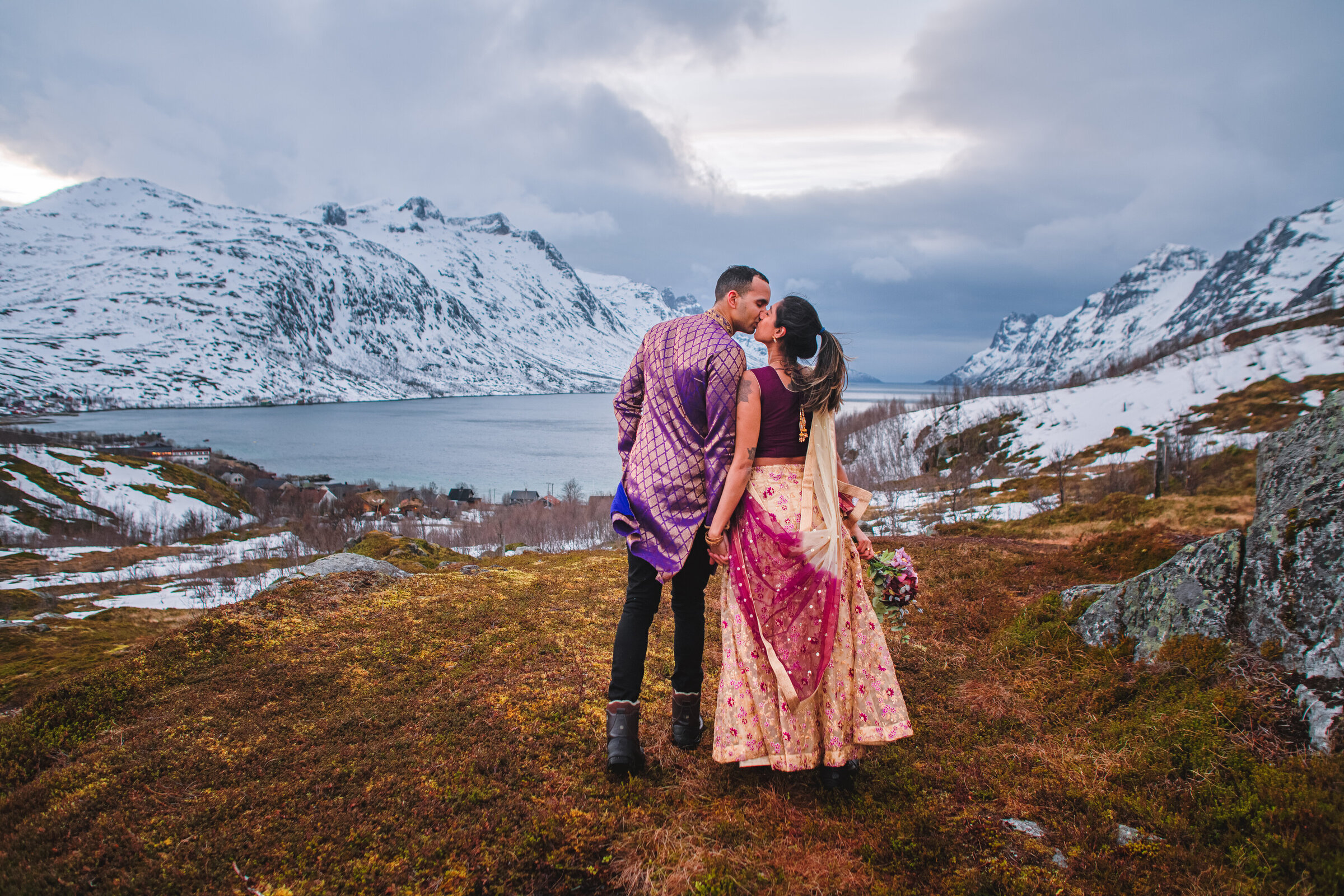 A couple in traditional Indian attire shares an  intimate kiss during their elopement, on a fjord overlook in Tromsø, Norway. They stand on a moss-covered mountainside, amidst a majestic, snow-capped mountain landscape. The man, dressed in a purple kurta with golden designs, gently kisses the woman, who is turned towards him wearing an ornate burgundy, pink and gold lehenga, with her back to the camera, both complementing the wild beauty of the Norwegian landscape. The sky above is filled with the dramatic February snow clouds and the soft glow of afternoon light, adding a touch of magic to the scene.