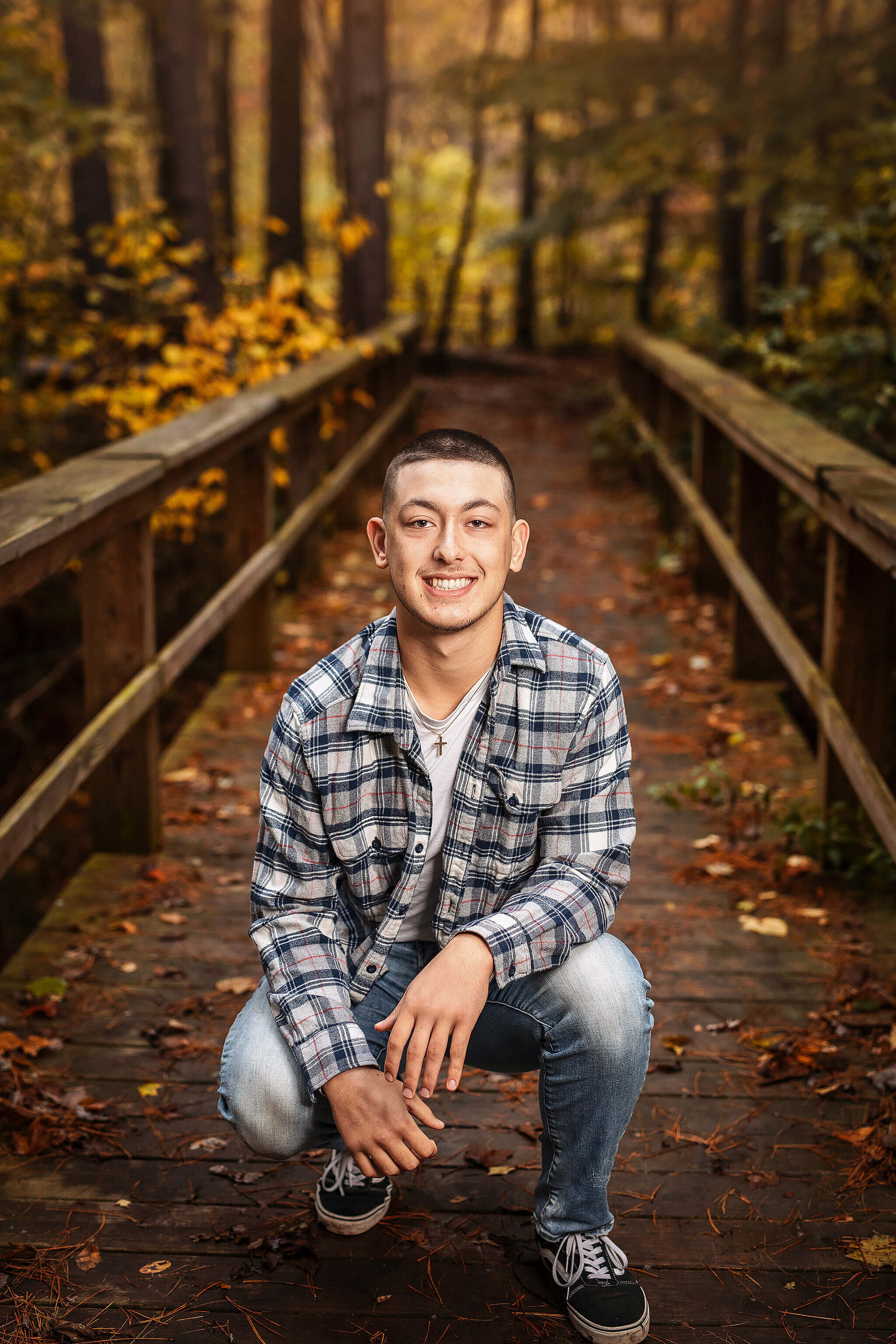 high school senior boy wearing a plaid shirt with jeans squatting on bridge