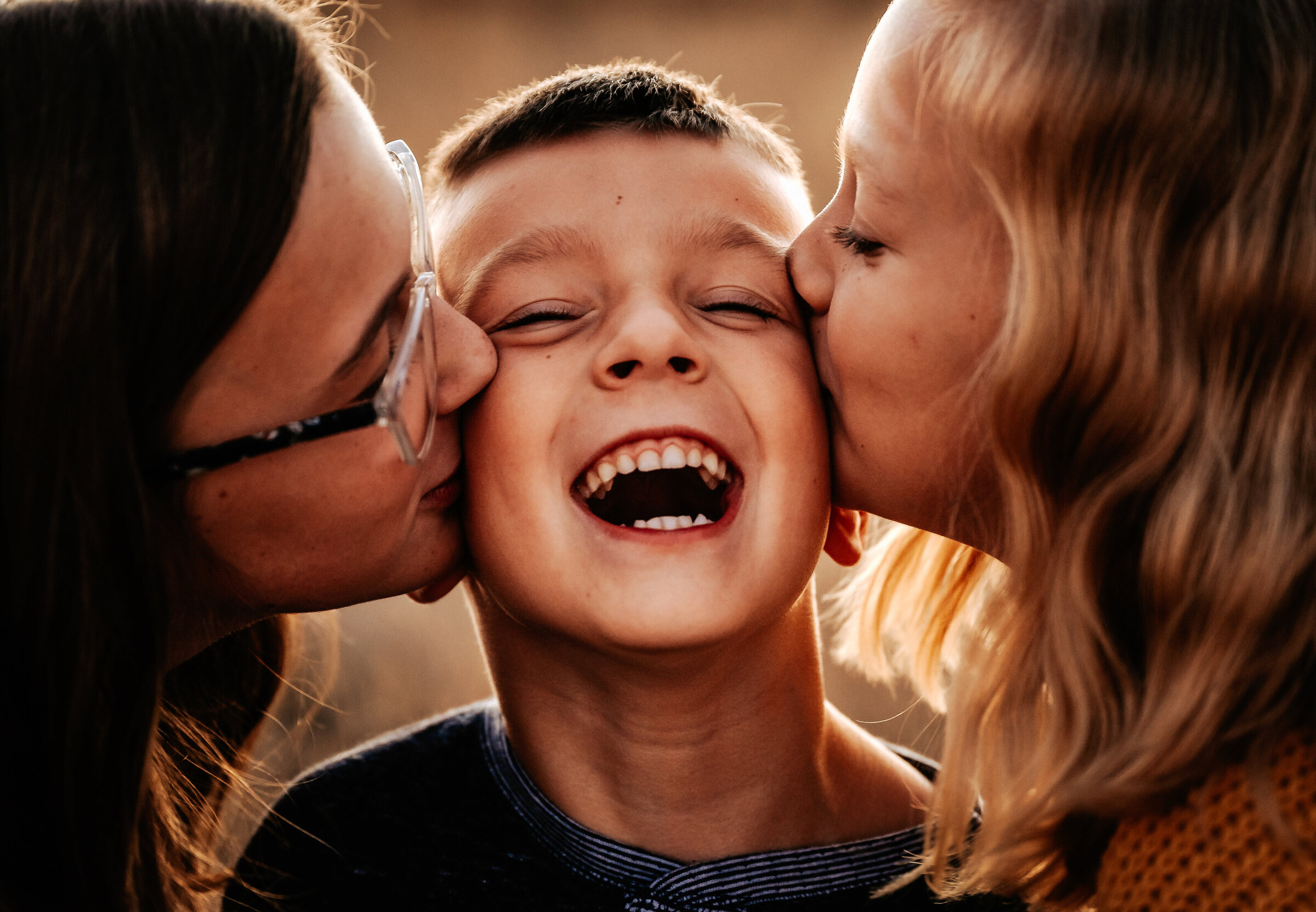 Sisters kiss younger brother on the cheeks while he laughs