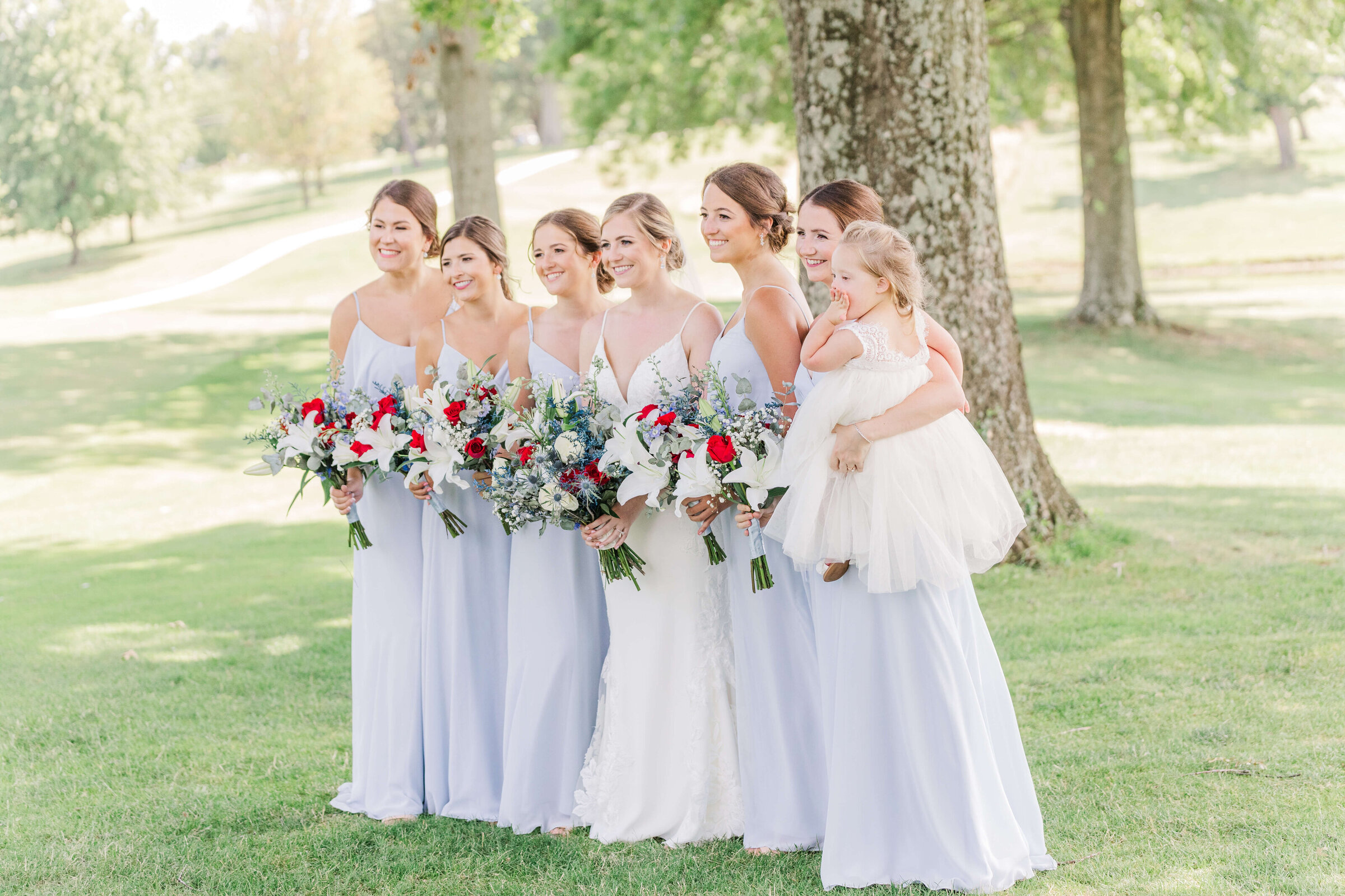 A bride and bridesmaids stand under a tree smiling at the camera. They are wearing light blue and the bride is holding a red and white bouquet.