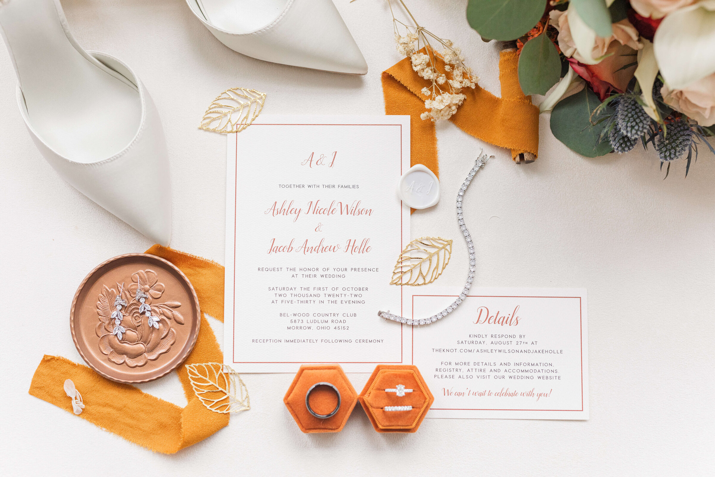 A wedding detail flat lay with an invitation suite, orange ring box with wedding rings, a small brown dish with earrings on it, white wedding shoes, orange ribbon and some flowers