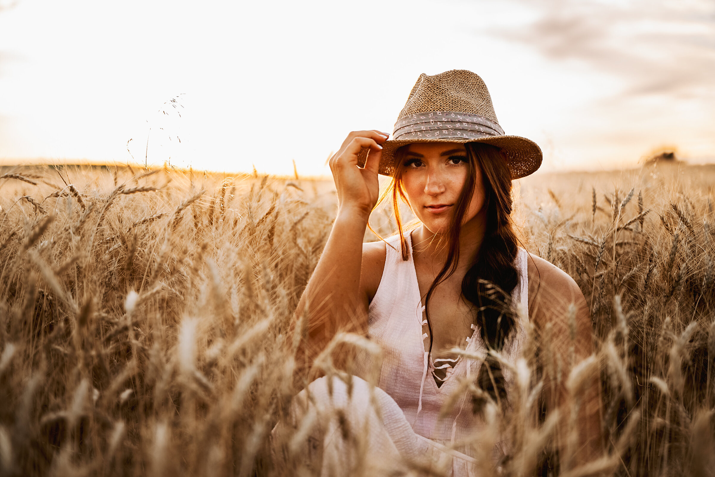 girl sits in wheat field and looks seriously at the camera while holding hat