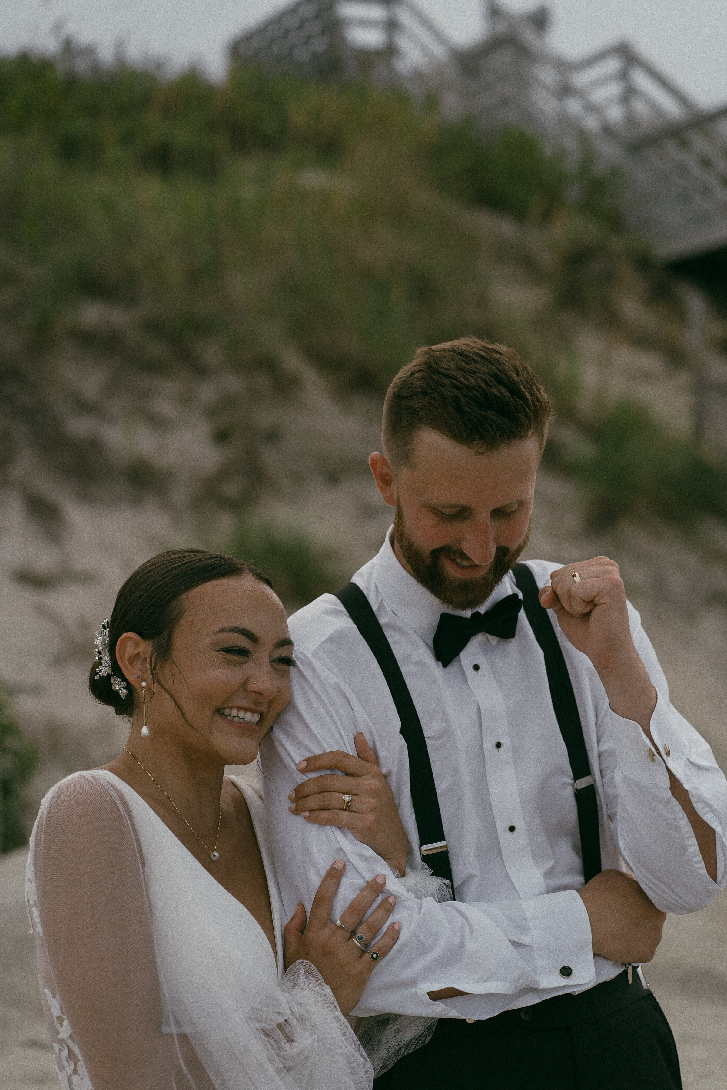 Groom adjusting his bow tie with bride laughing beside him on the beach.