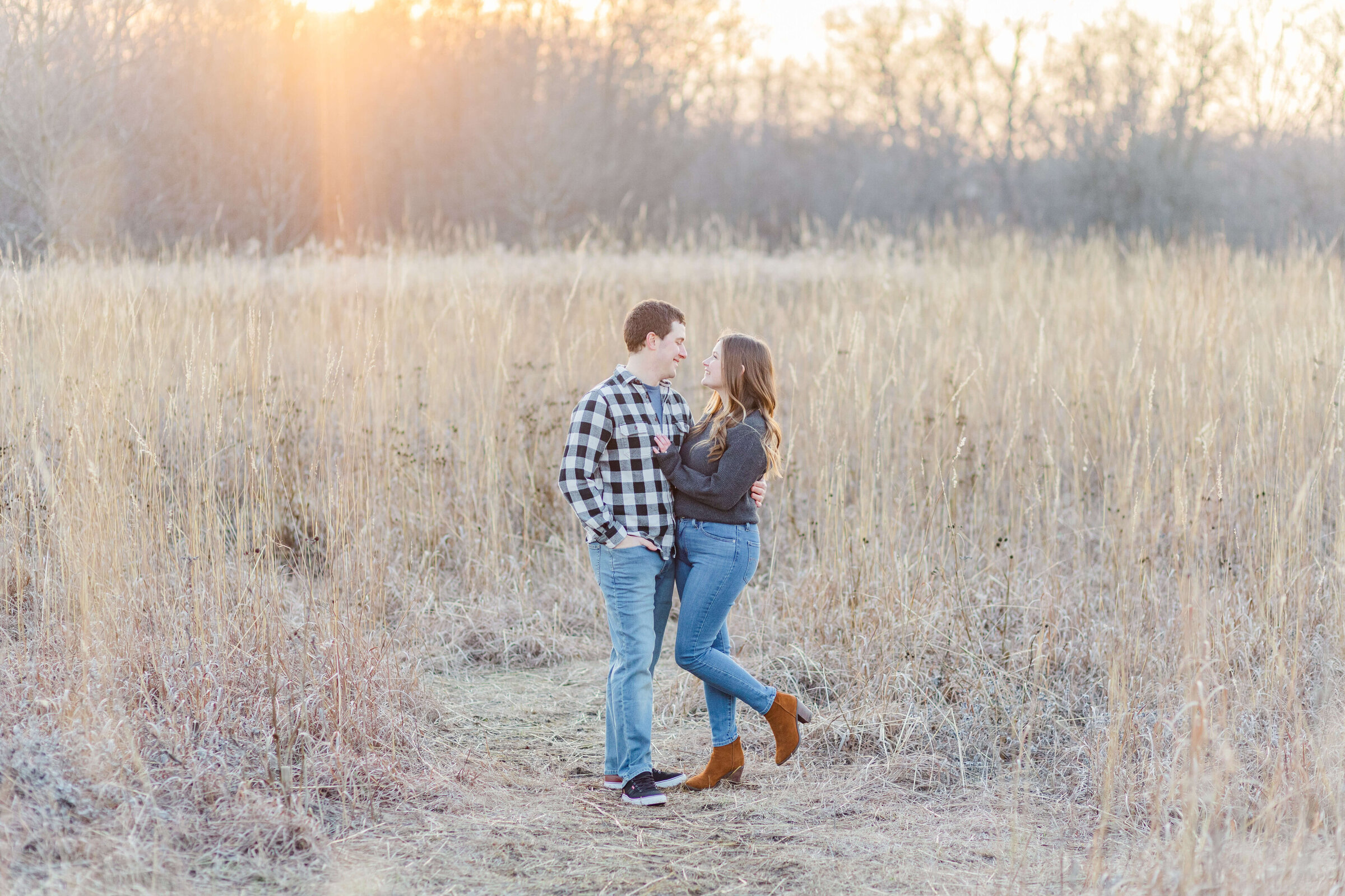 A couple is in in a field with the sun glowing on them. They're both in jeans and grey shirts