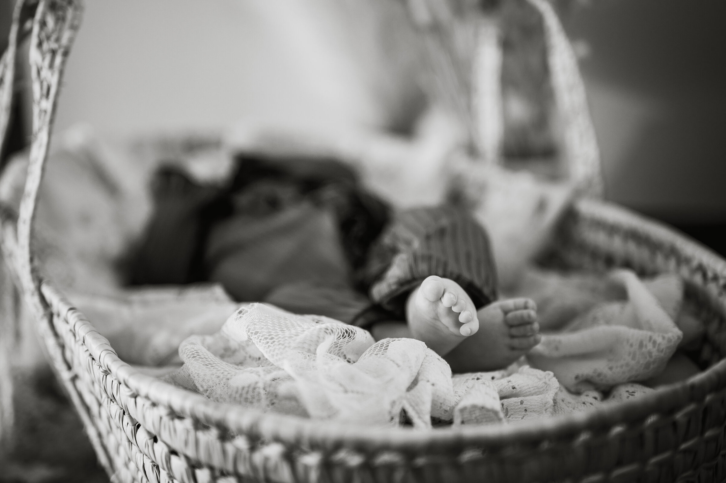 Black and white image of baby's toes while baby lays in a moses basket filed with linens and canvas scraps