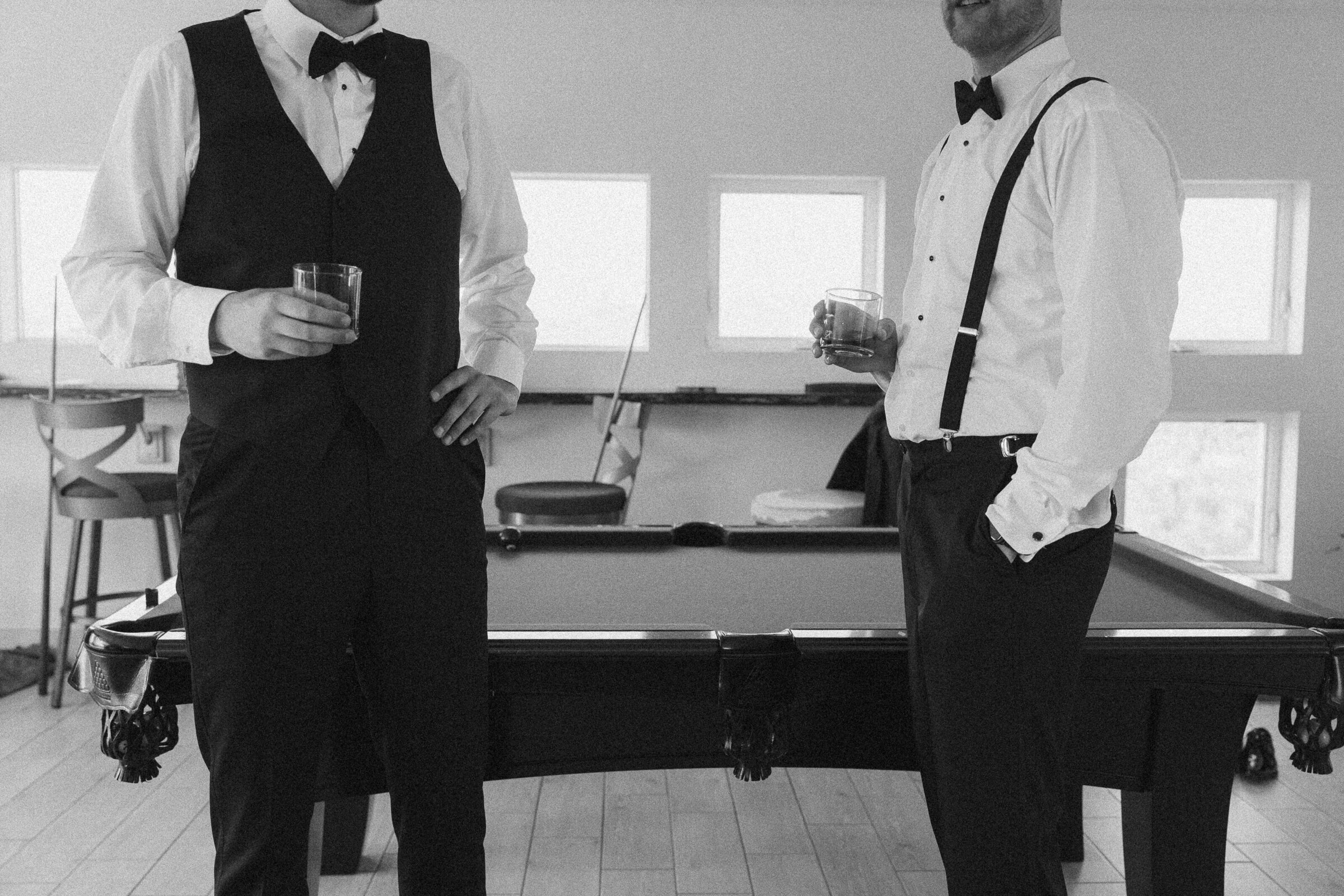 Two men in tuxedos with bow ties holding whiskey glasses near a pool table.