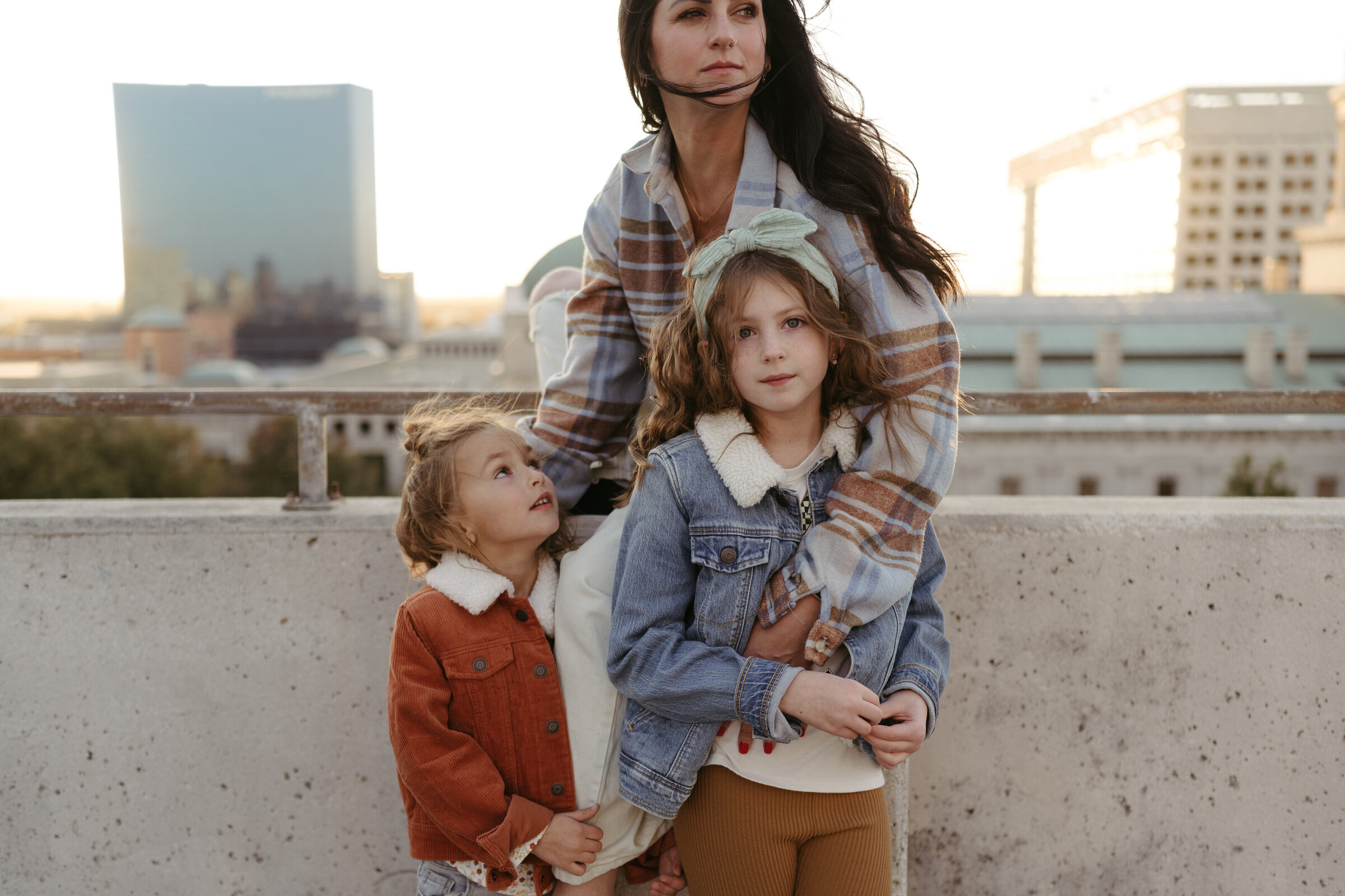 A mom and two young girls pose on a parking garage at sunset while the wind blows their hair
