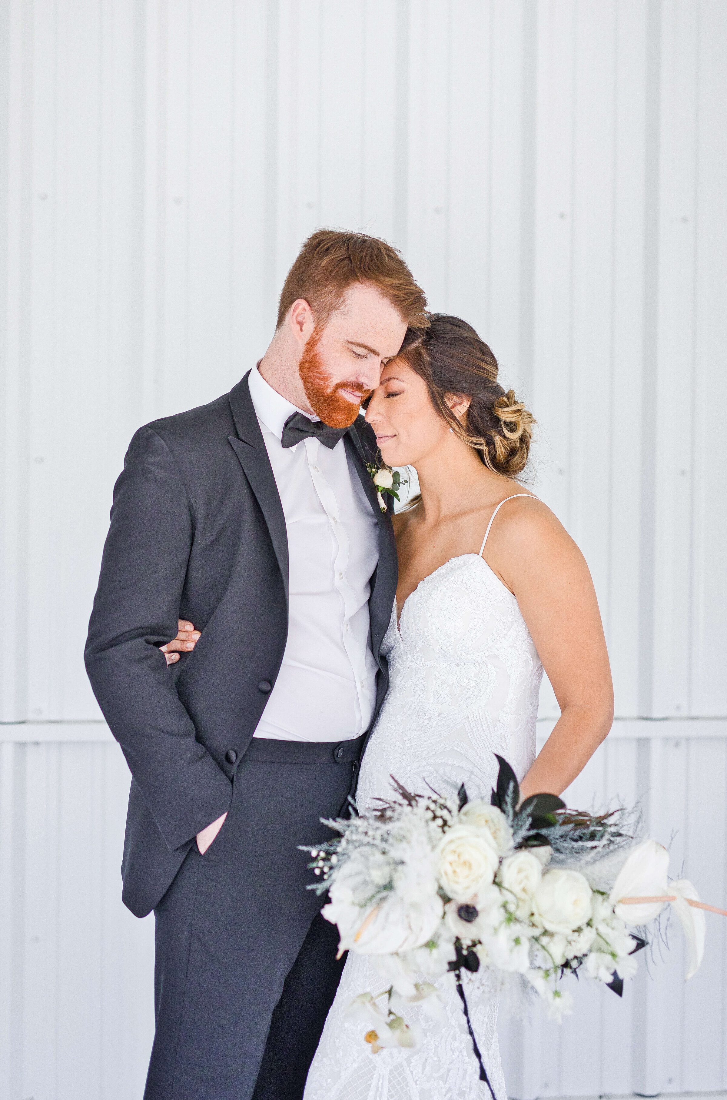 A groom and bride stand next to each other with their arms around one another with their eyes closed. She is hold a big white bouquet.