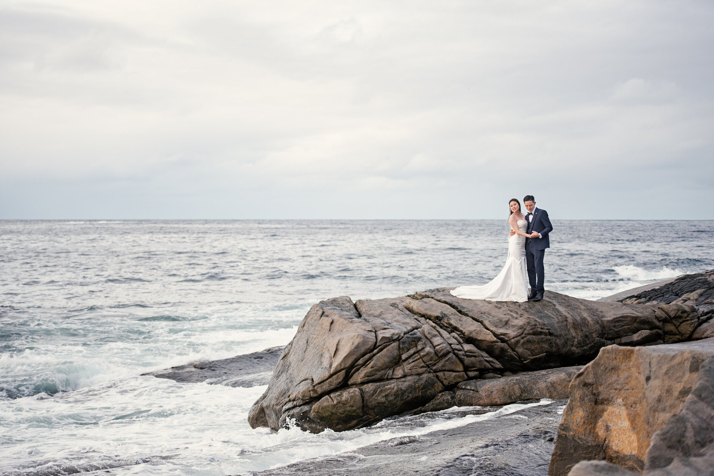 A couple stands closely embraced on a large rock by the sea in Senja, Norway, with waves gently breaking against the rocky shore around them. The vast ocean behind them meets a cloudy sky at the horizon, reflecting the serene and isolated setting of their elopement.