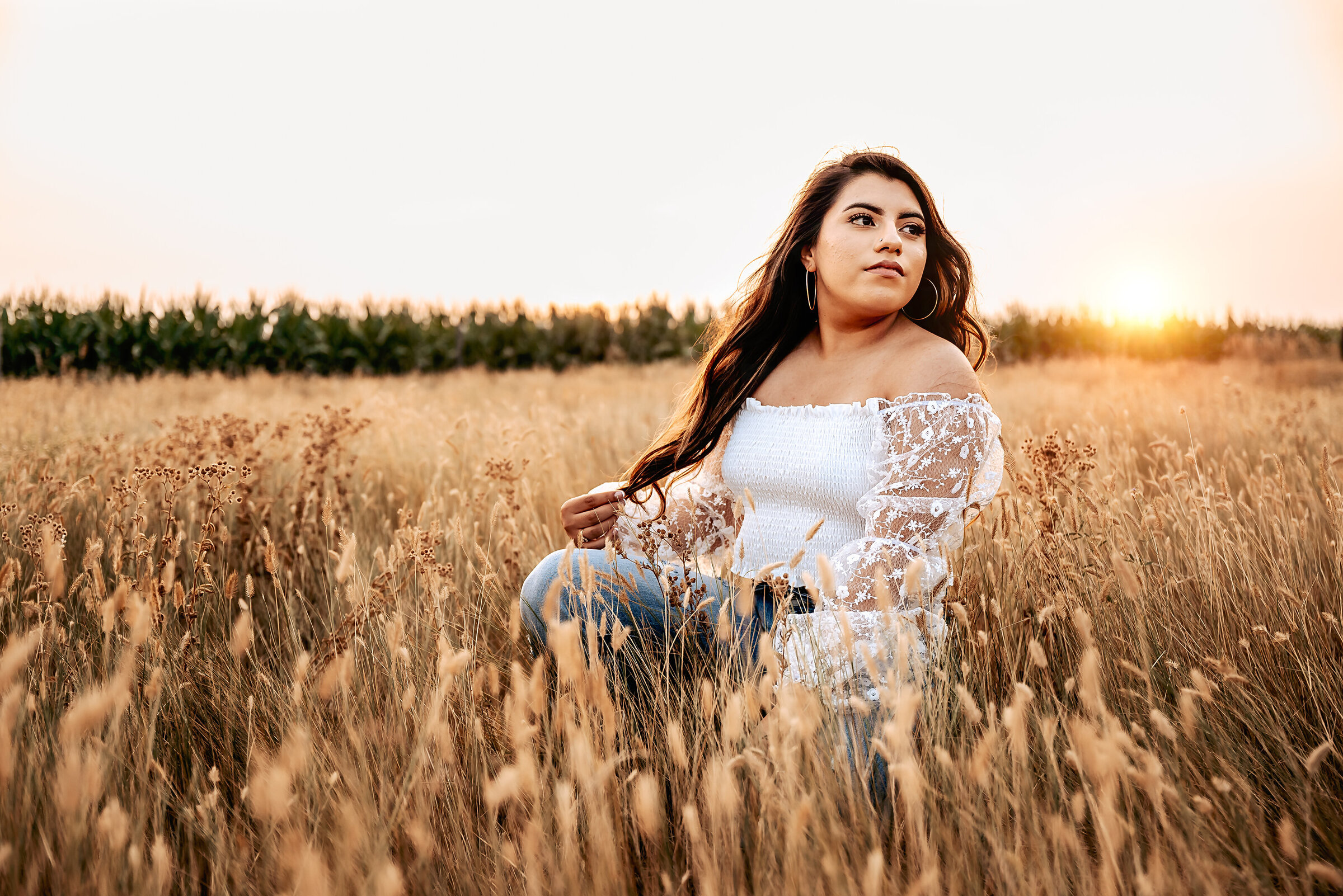 Girl kneels in open field for senior pictures and plays with her hair