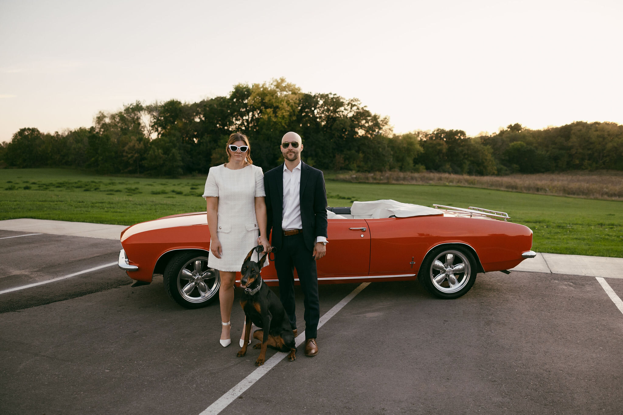 Future bride and groom standing in front of a red classic convertible with their dog in between them, engagement photos captured by Morgan Ashley Lynn Photography in Madison, WI