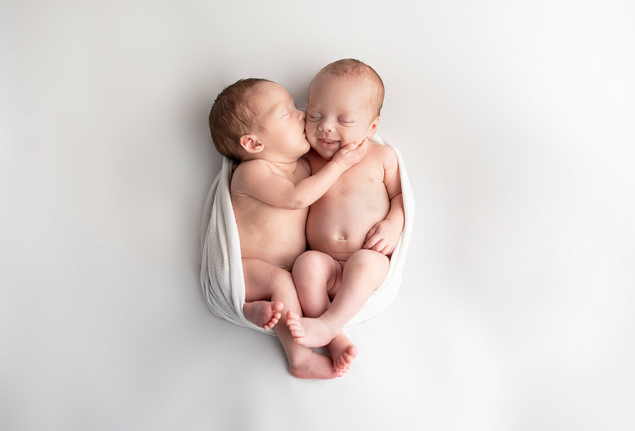 newborn twins snuggled together on a white blanket while one twin gives the other twin a kiss on the cheek