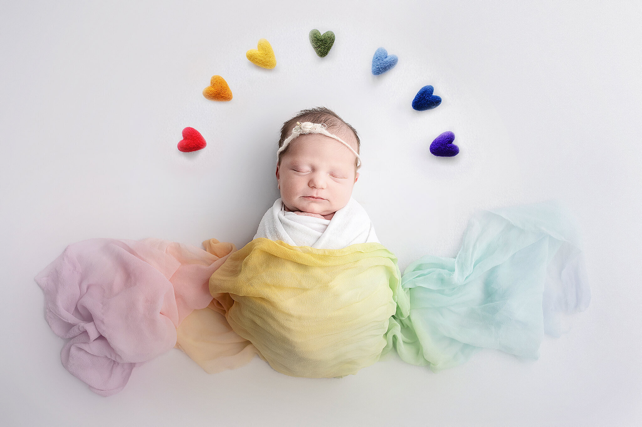 newborn rainbow baby wrapped in rainbow wrap with rainbow colored hearts above her head in a  rainbow shape.