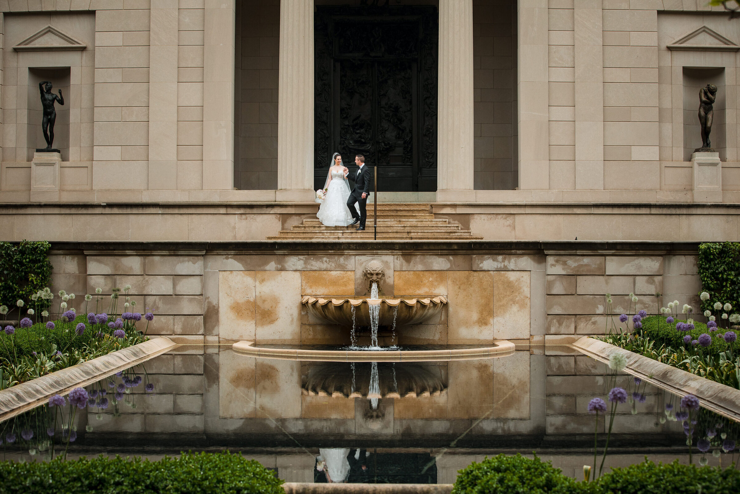 The groom is walking the bride down the steps at Rodin Museum wedding in Philadelphia.  Their reflection can be seen in the beautiful pool.
