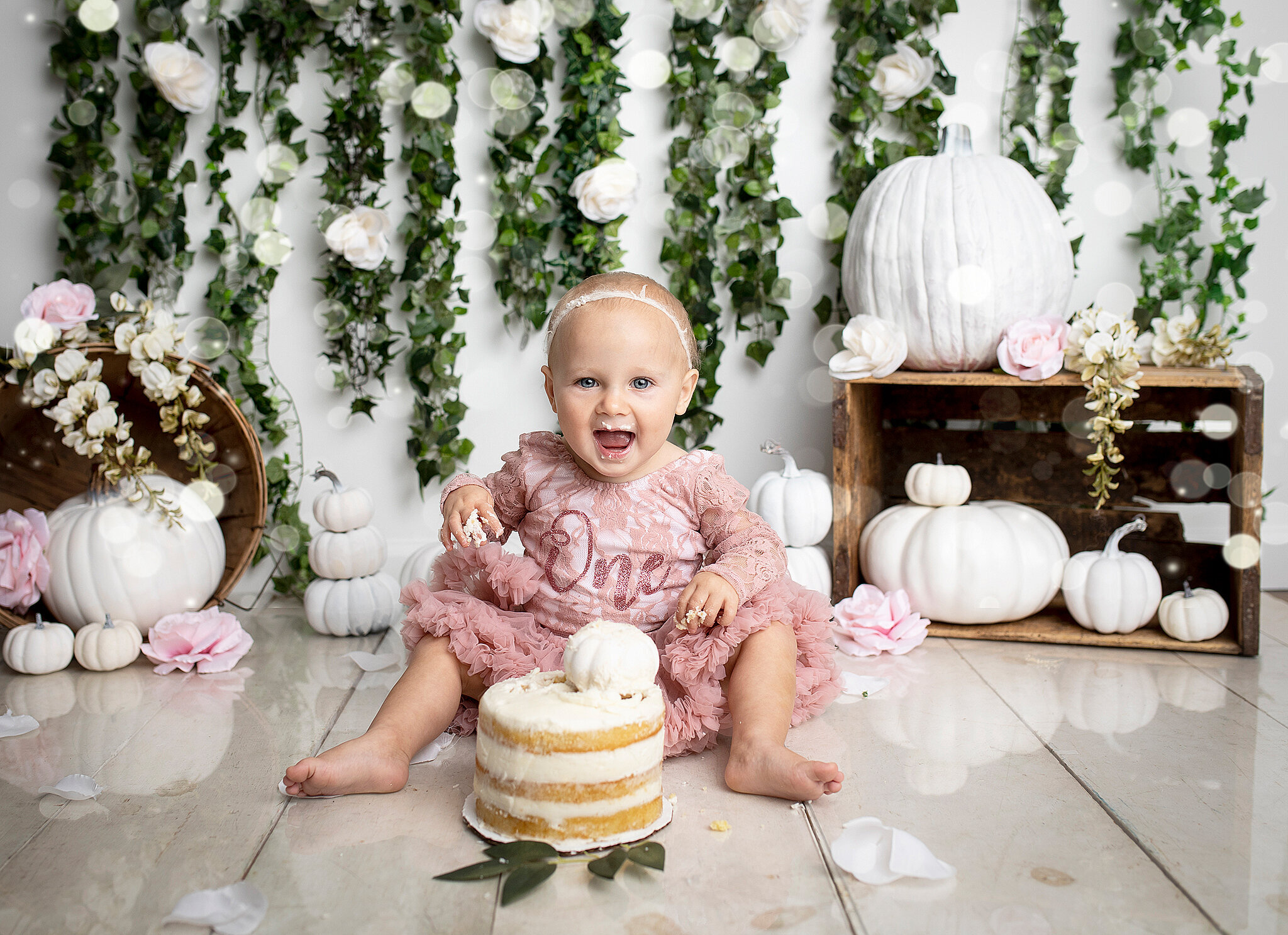 baby smashing white cake in pink dress, white pumpkins and greenery in the background