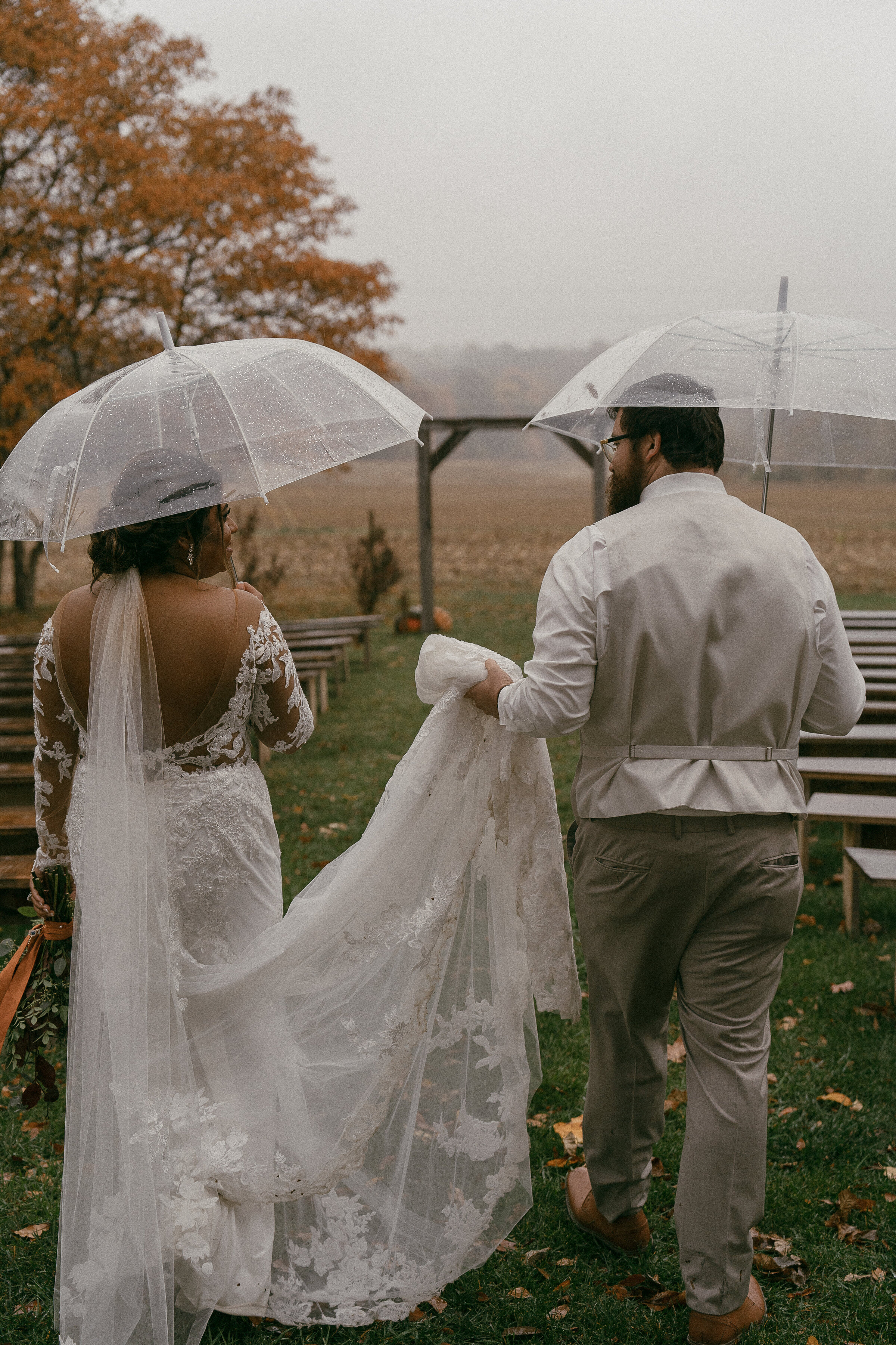 Bride and groom walking under clear umbrellas on a rainy day.