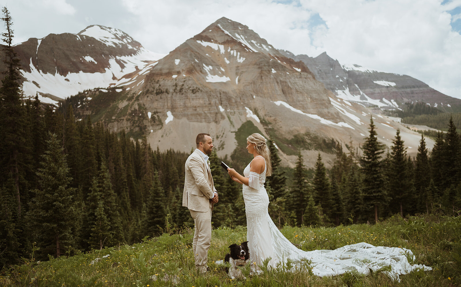 The couple is facing each other with their dog in between them as they read their vows in a valley. It is summertime and very warm. The bride is wearing a dress with patterned lace, and the groom is wearing a tan suit. They are wiping away tears as they finish their elopement. The couple decided to elope in Colorado, and they hiked and off roaded on their elopement day. They included their dog in their elopement, which is a border Collie. The mountains are directly behind them and are framing them in this image. there is still snow on them, and there are wildflowers. If you want to find wildflowers on your elopement, plan to elope in the early summer.