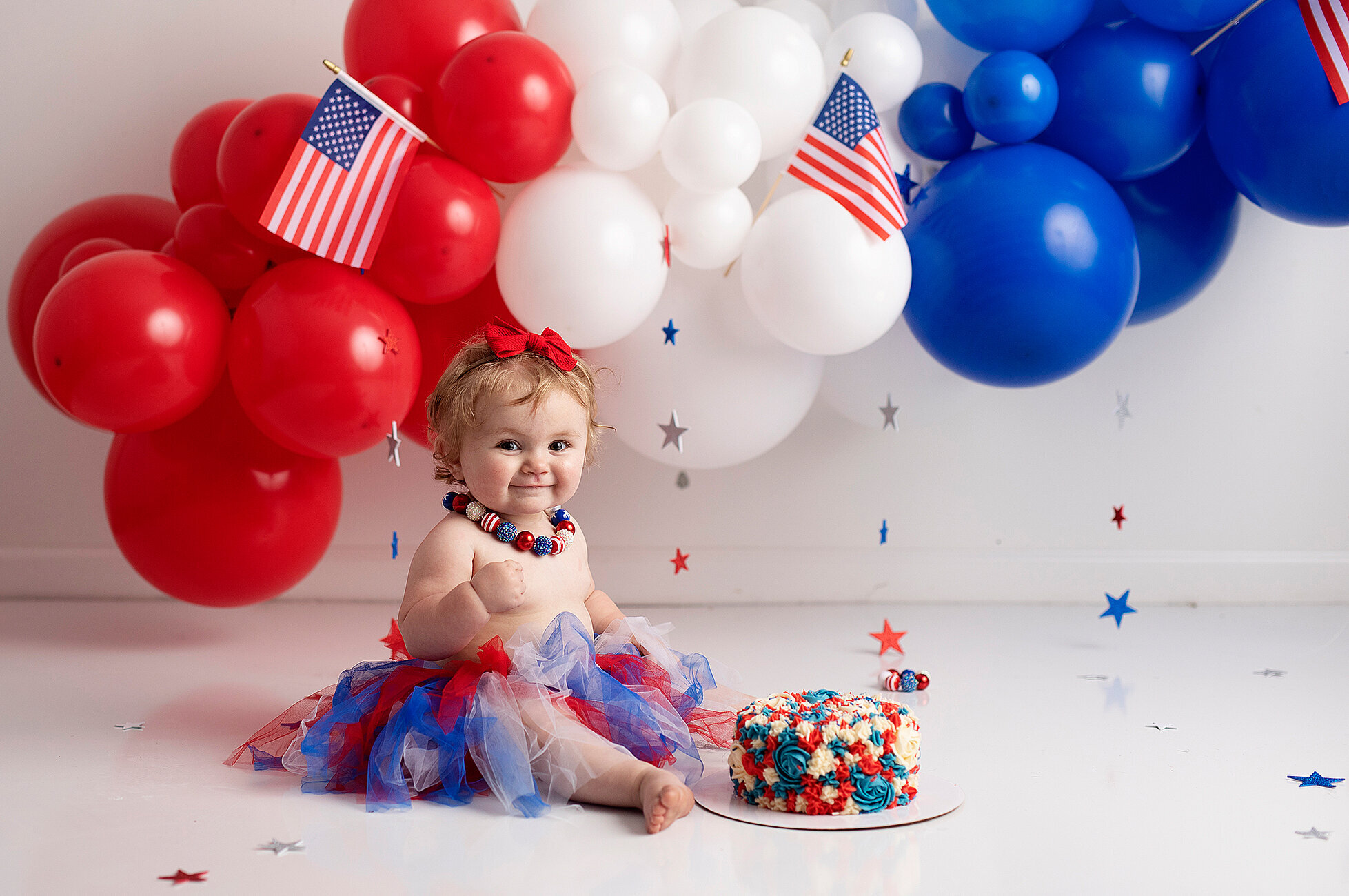 One year sitting with red, white and blue cake and red, white, blue balloons in the background
