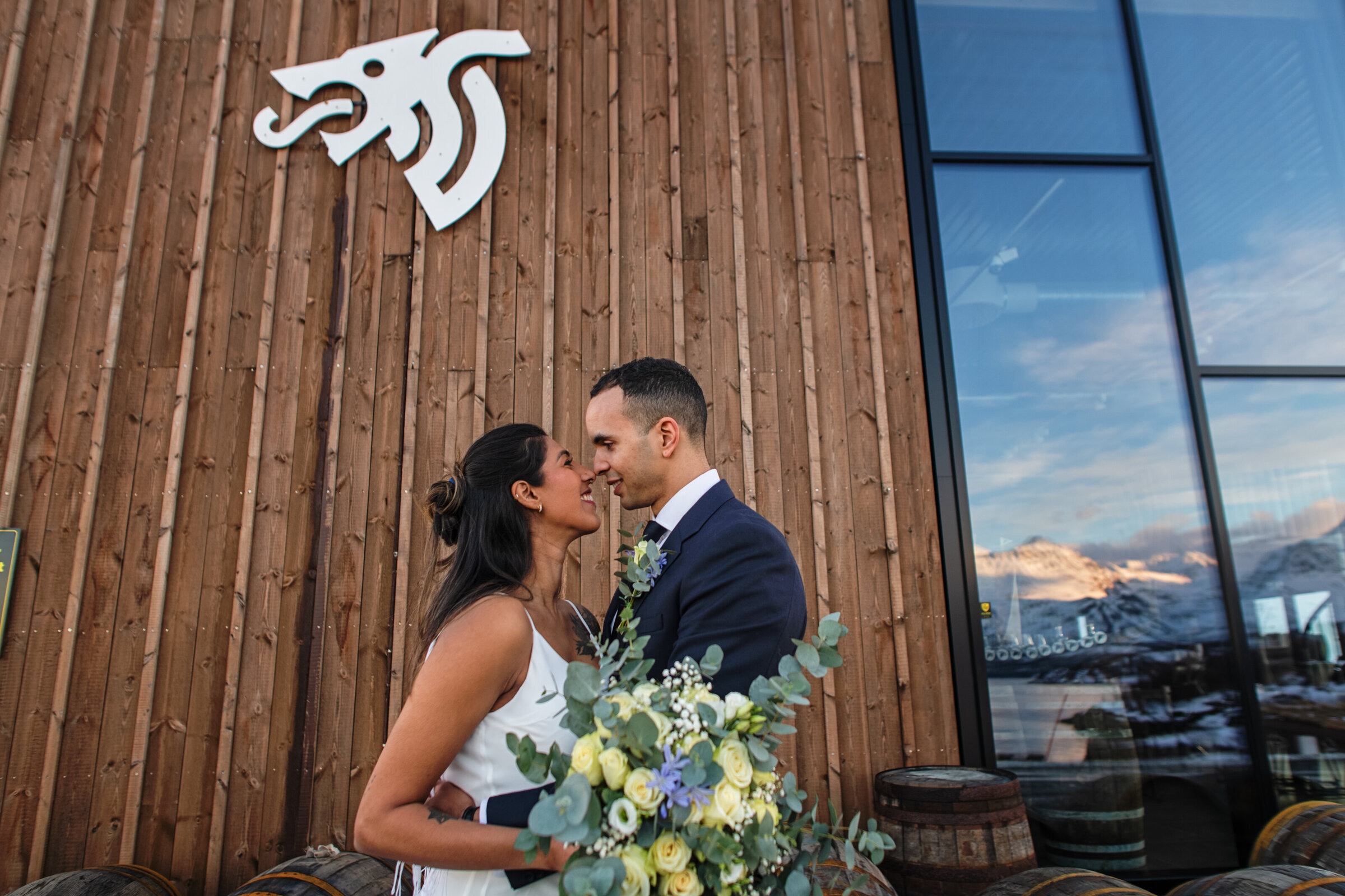 A close embrace between the bride and groom outside Aurora spirit distillery in Lyngen, Norway. Highlighting their unique elopement location, the bride also holds a bouquet of locally sourced and designed flowers, showcasing the vendor connections available with their elopement planner.