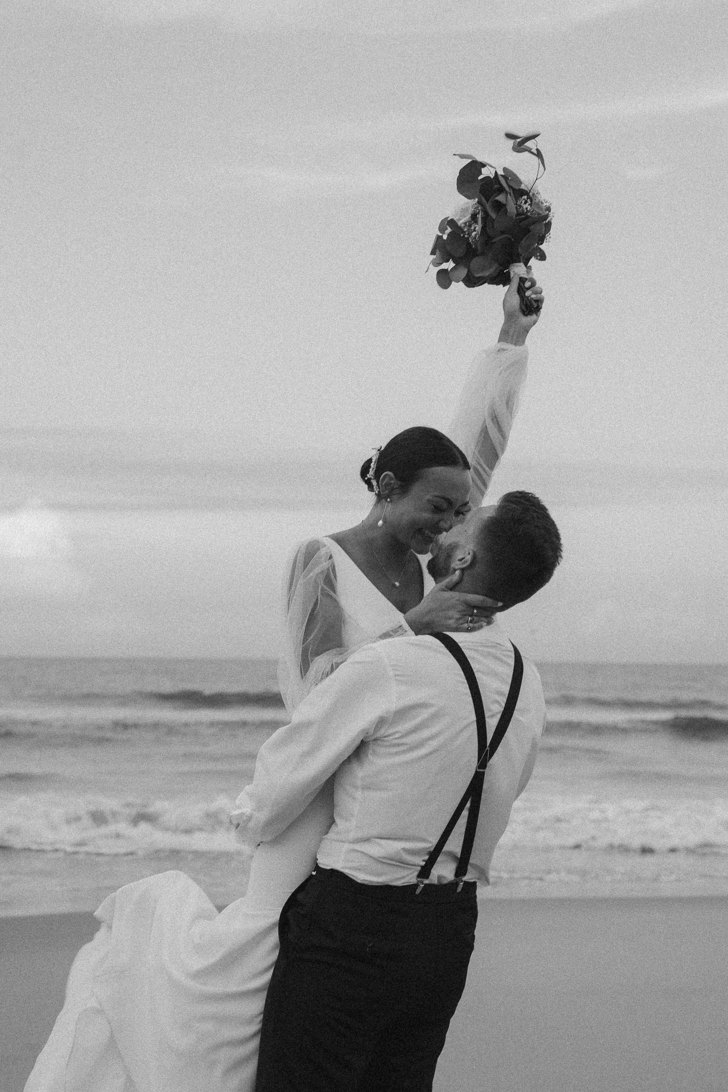 Bride lifted in the air by the groom on the beach, with a bouquet tossed in the air.