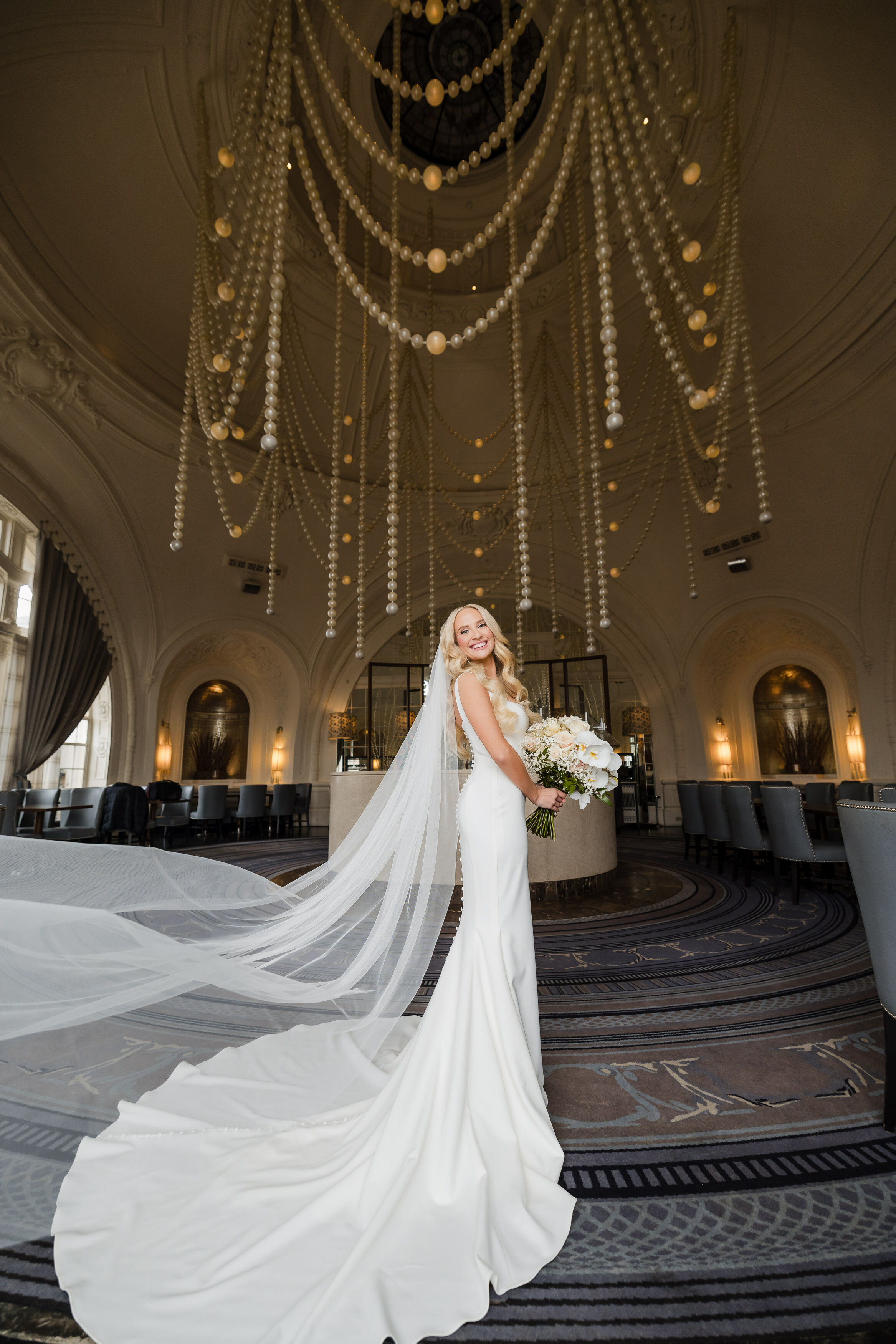 A wide angle bridal portrait of an elegant blond bride with a flowing veil under the pearl chandelier at the XIX at Bellevue on her wedding day in Philadelphia