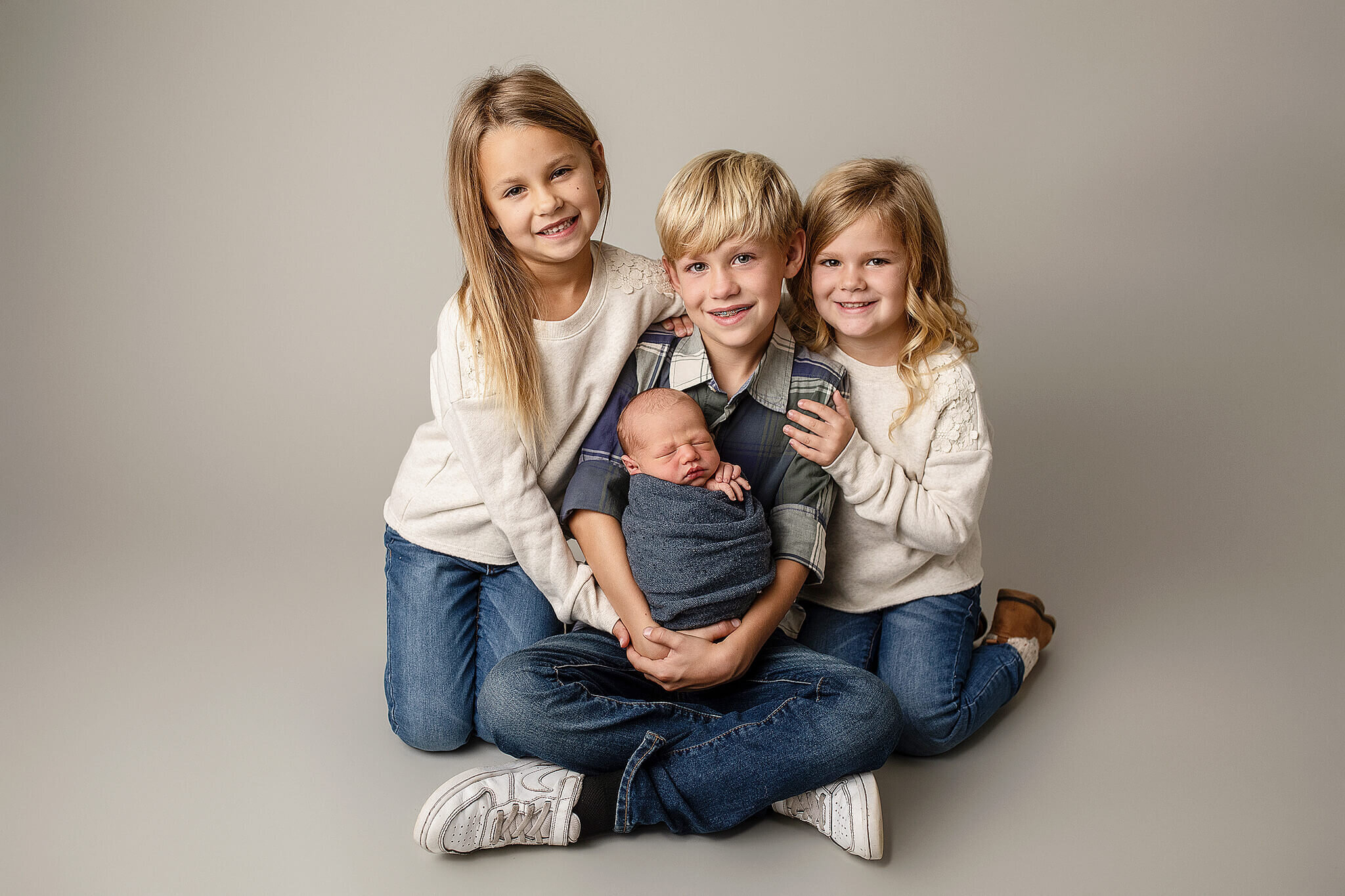 Three older siblings with the newborn brother.  Kids sitting on grey paper background and big brother is holding baby brother who is wrapped in blue wrap.