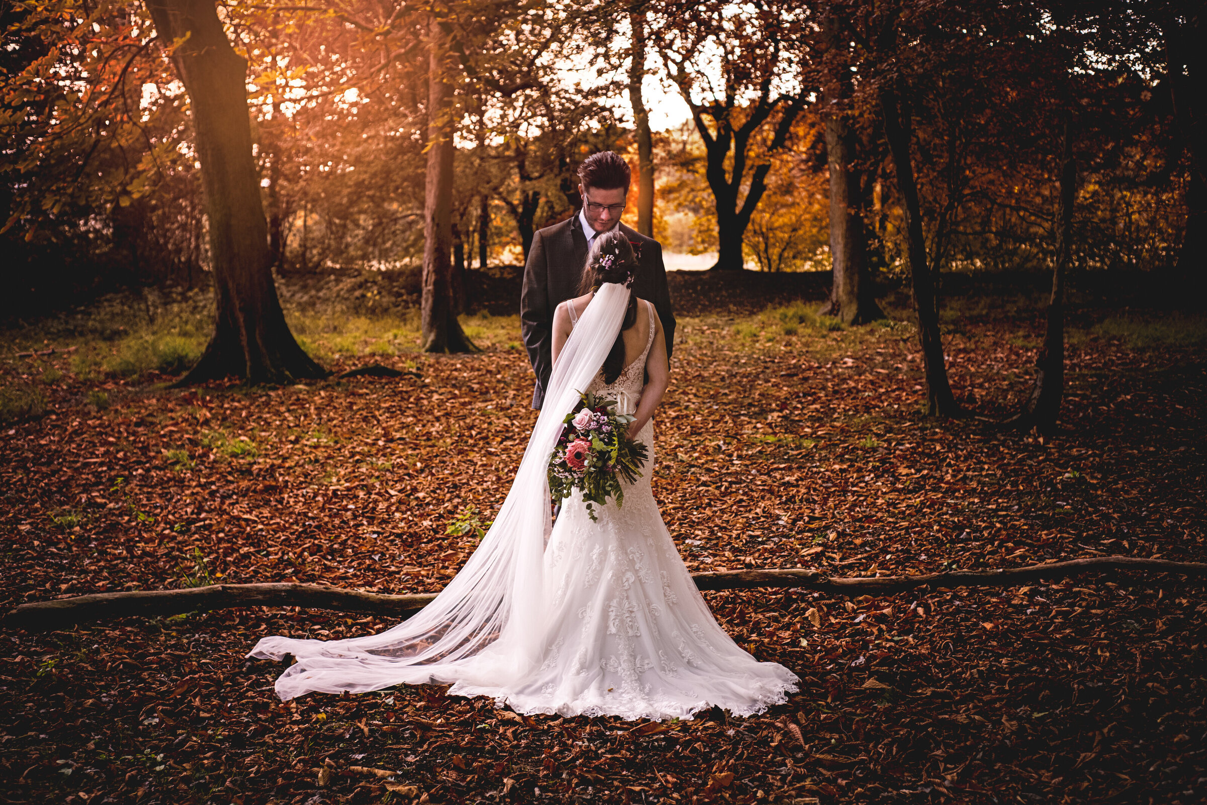 In a tranquil autumn forest, a newly eloped couple embraces in a quiet moment, surrounded by fallen leaves. The bride, in an exquisite gown with a flowing veil, and the groom, in a classic suit, capture the essence of their secluded and romantic celebration.