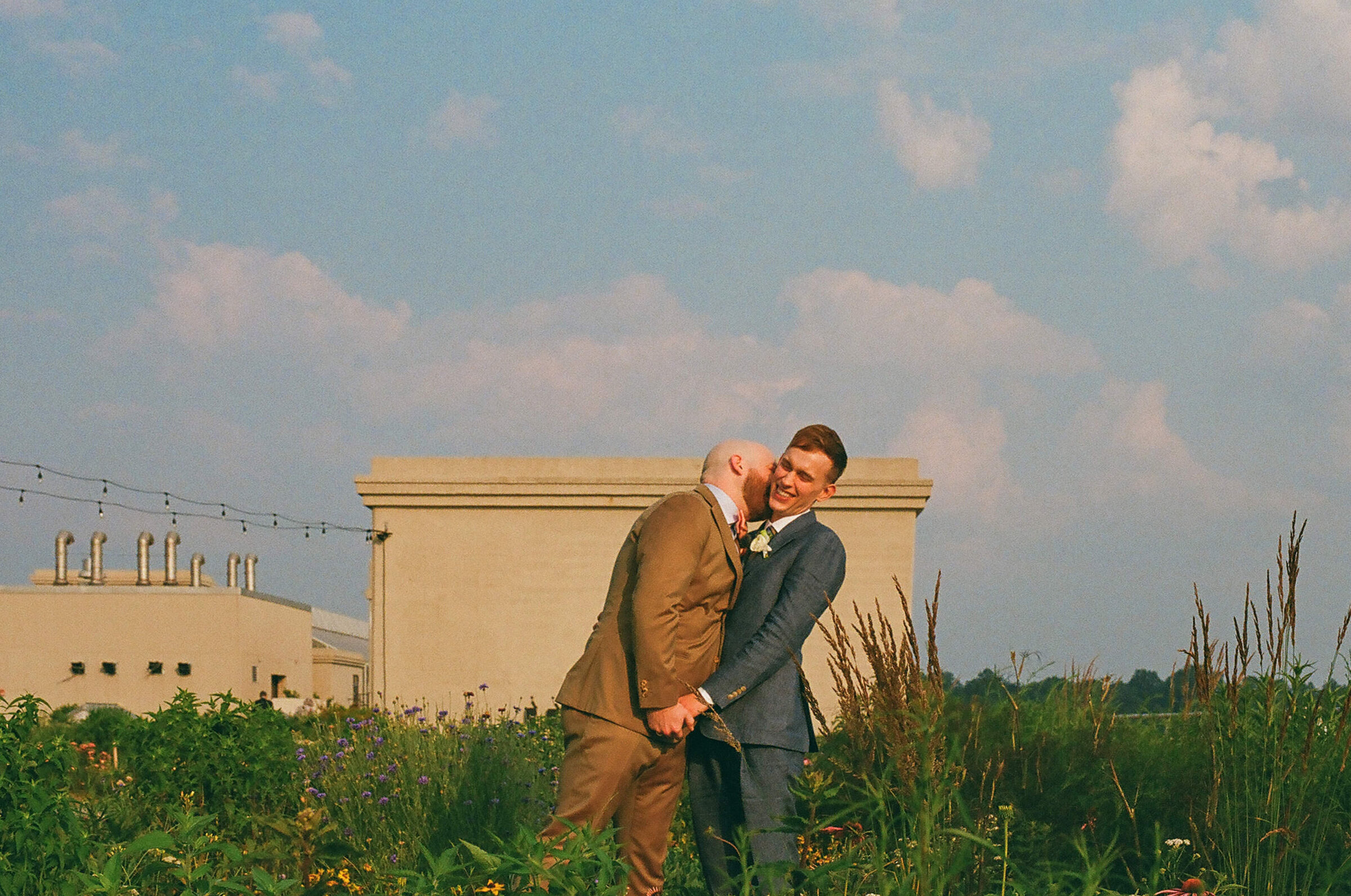 A person leaning in to kiss their partner's cheek in a small field.