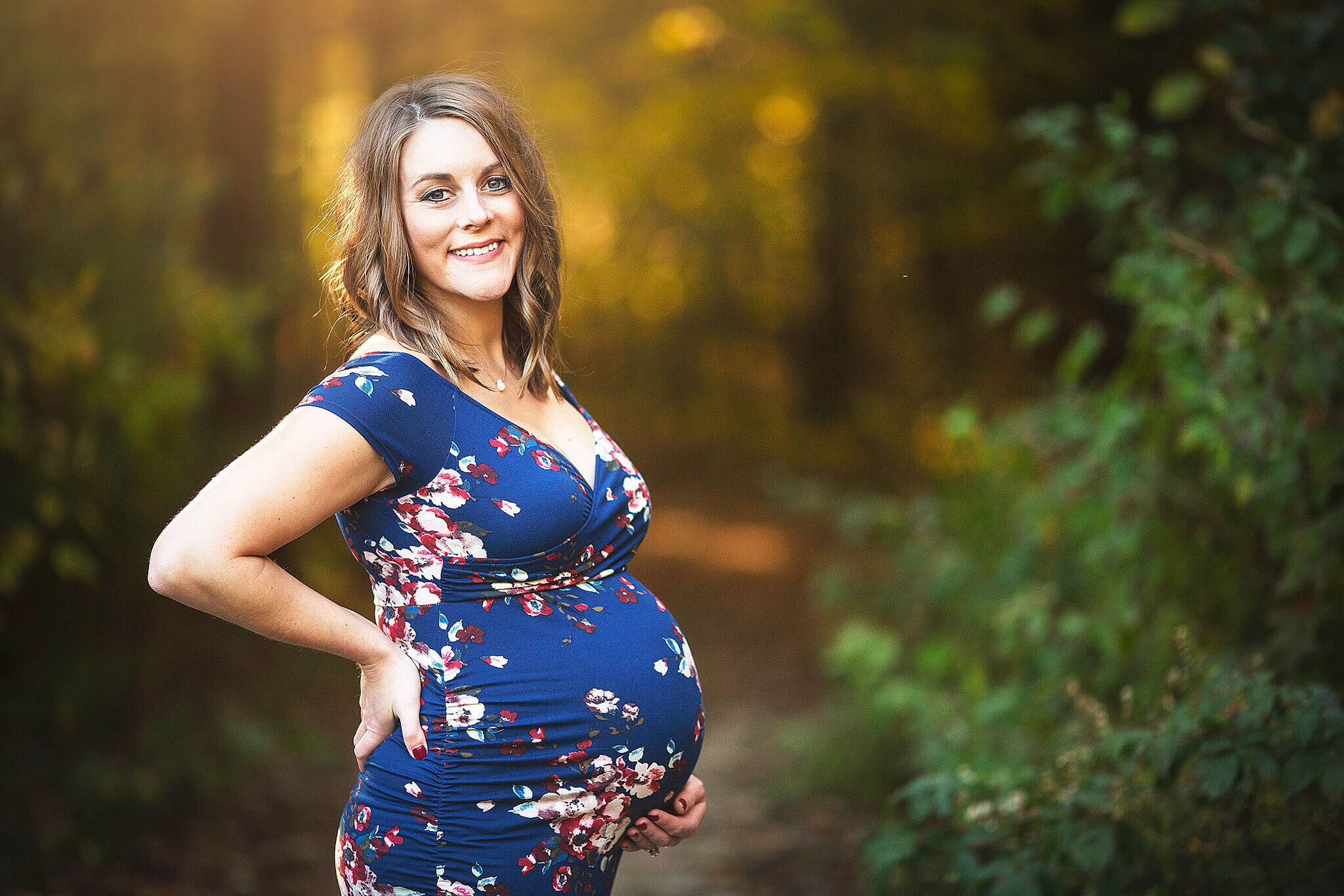 Pregnant women in blue dress with flowers on it, with one hands on her belly and one hand on her back, standing in front of a path in woods