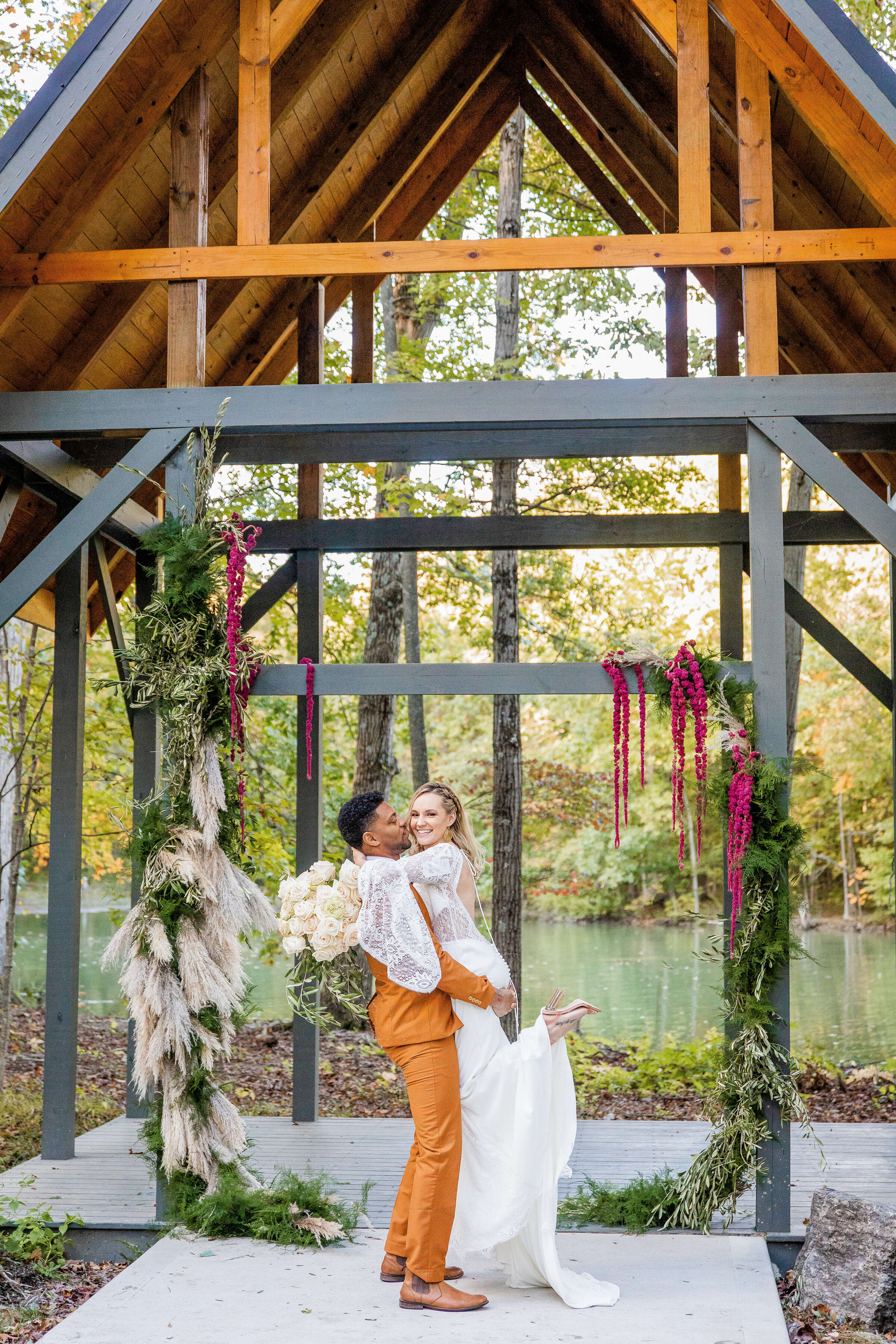 A groom in a dark orange suite picks up his bride as she smiles. They are standing under a gazebo on a lake.