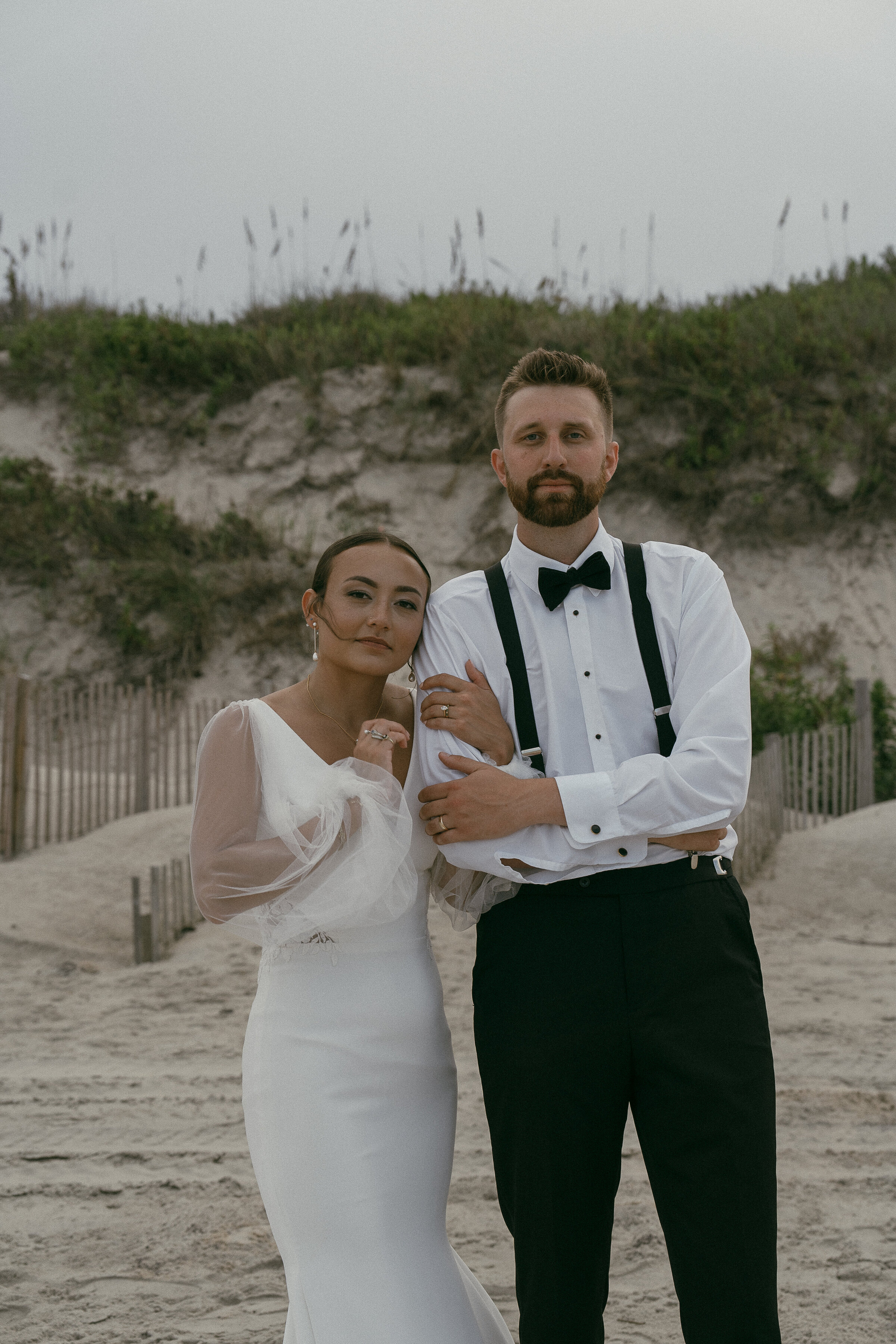 Bride and groom standing on the beach with serious expressions.