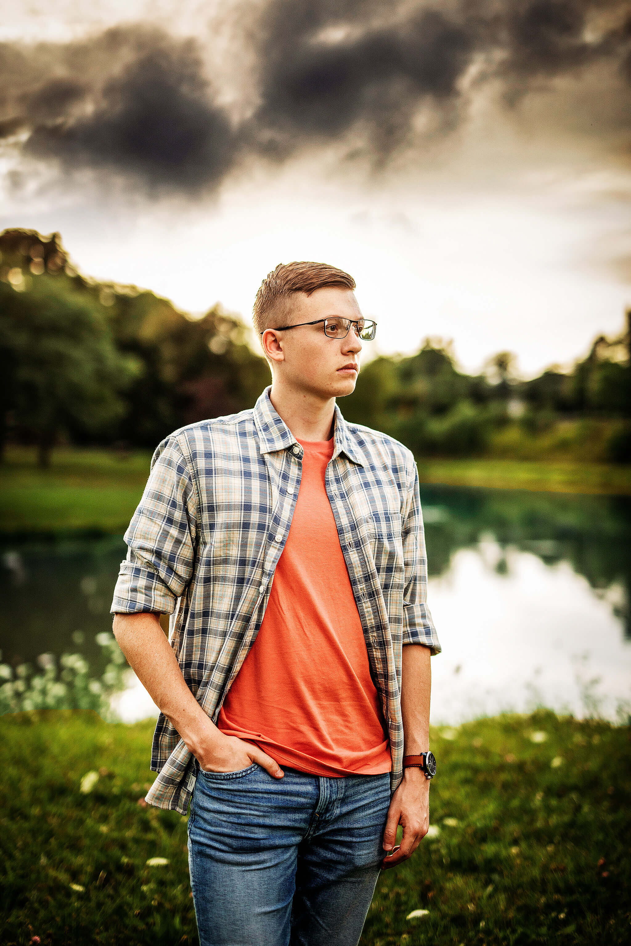 high school senior boy wearing orange shirt, jeans and a plaid button up standing in front of a pond in ashland ohio
