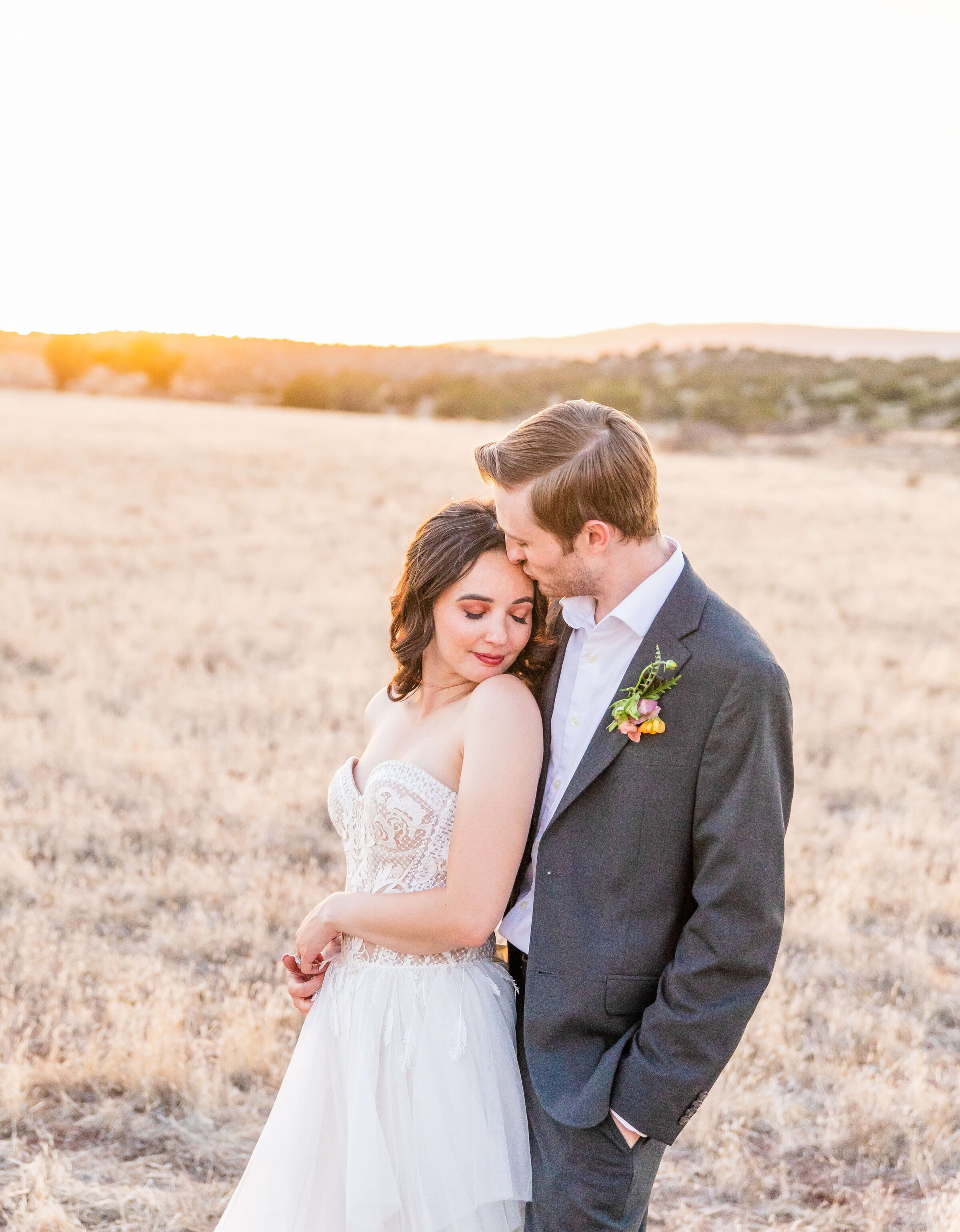 A groom kisses the forehead of his bride standing in the Sedona field as the sunsets