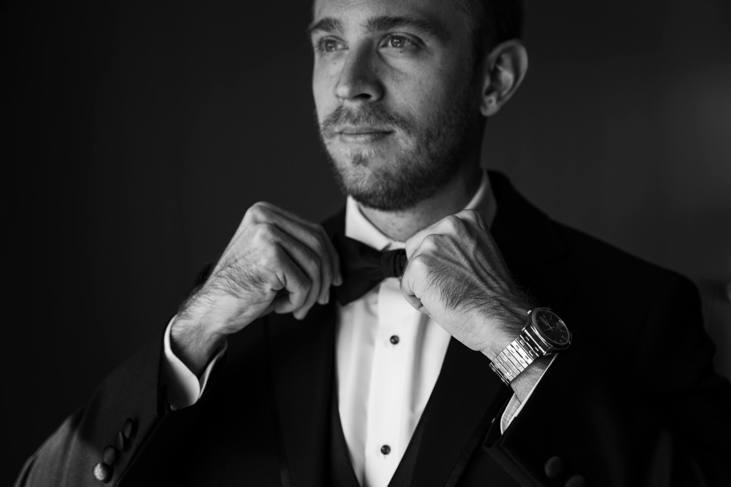 An up close black and white photograph of a groom straightening his bowtie