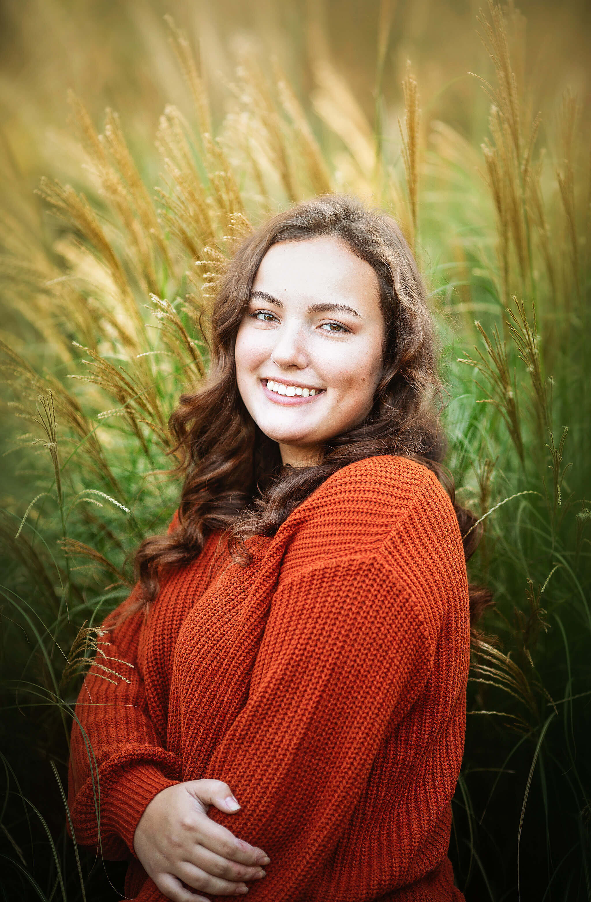 high school senior girl in orange sweater standing in front of tall green grass