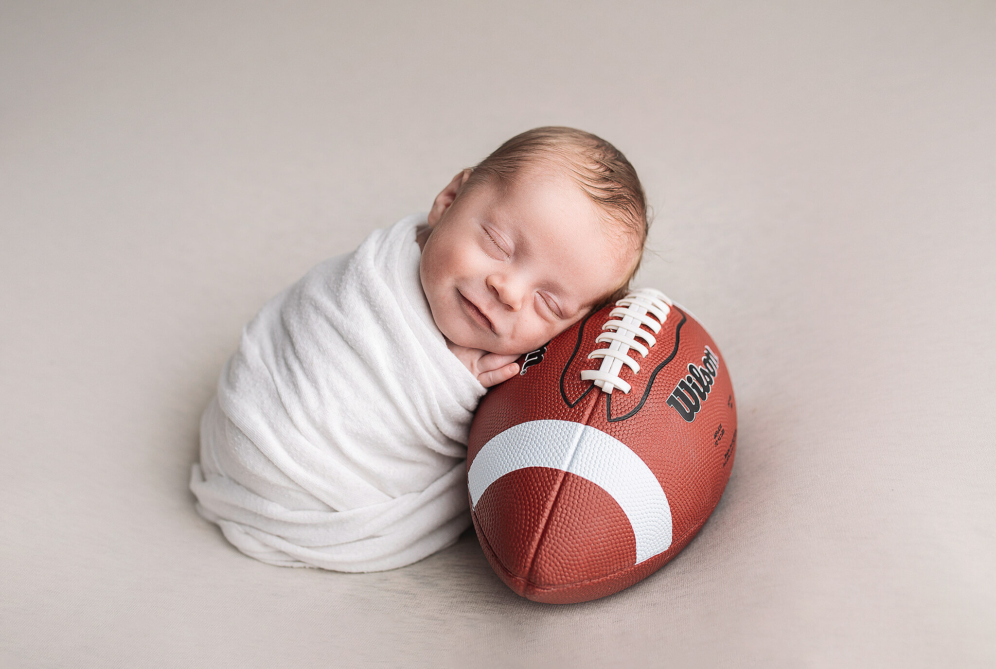 newborn baby boy on tan blanket, wrapped in white and leaning while smiling on  a football