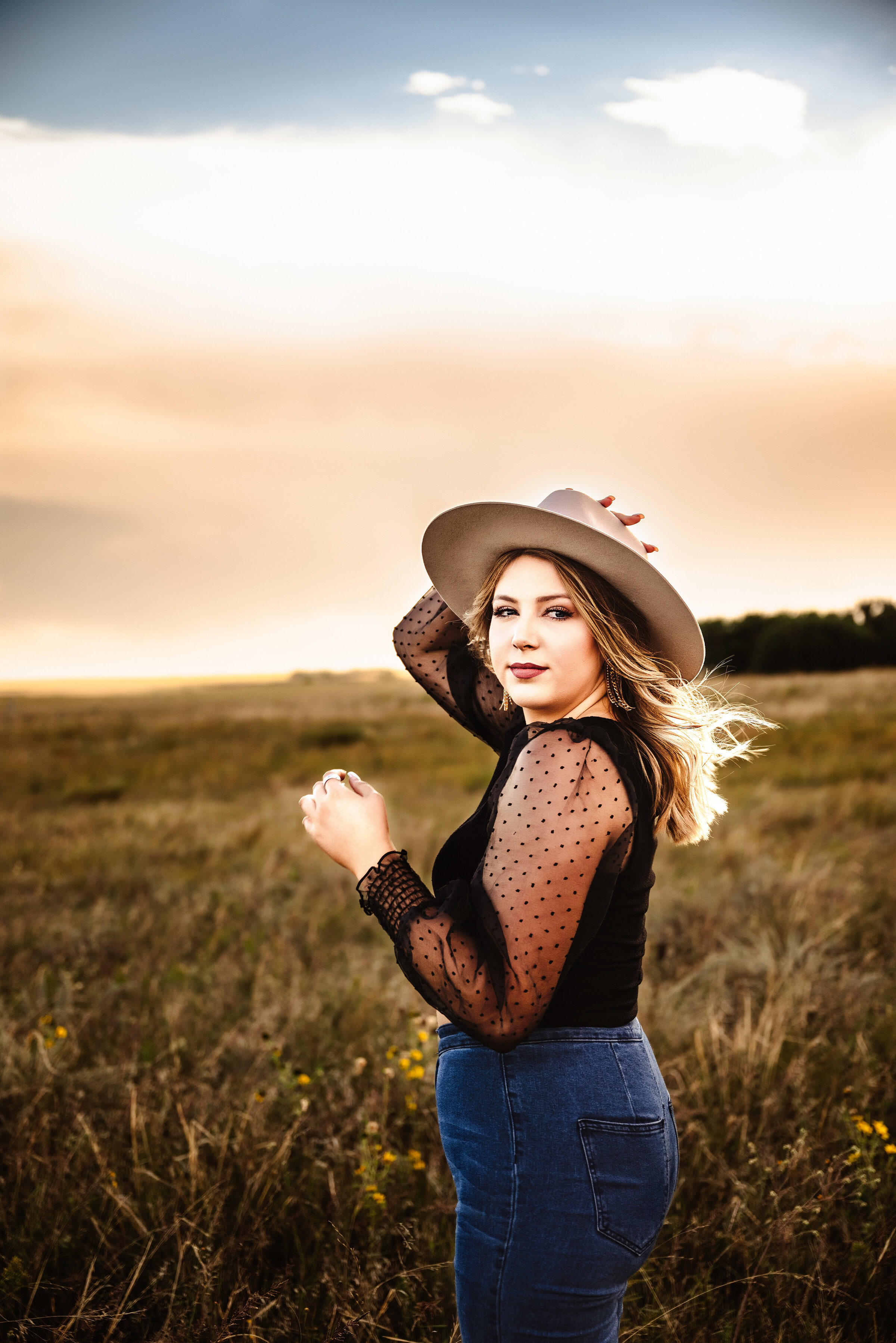 Senior girl holds onto hat while wind blows her hair in open field