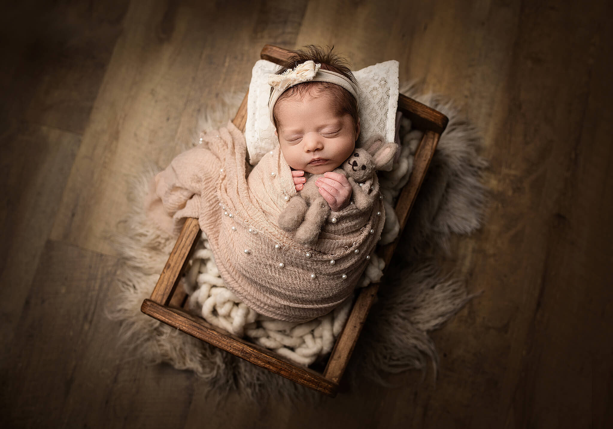 newborn baby girl wrapped in tan wrap with pearls, while lying in a littel brown cracdle and holding a little stuffed bunny.
