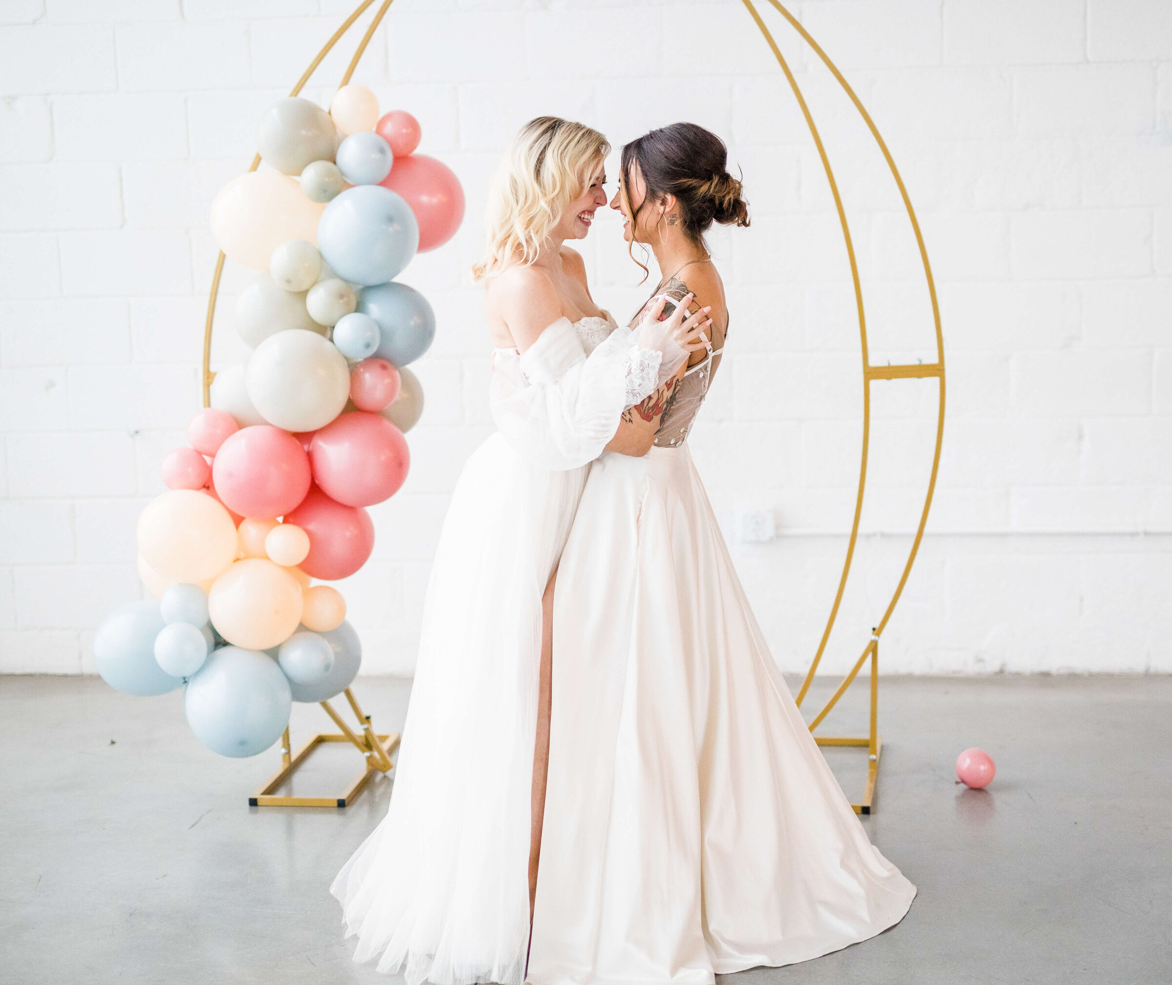 Two brides hug each other as they laugh. There is decor in back of them with rainbow balloons