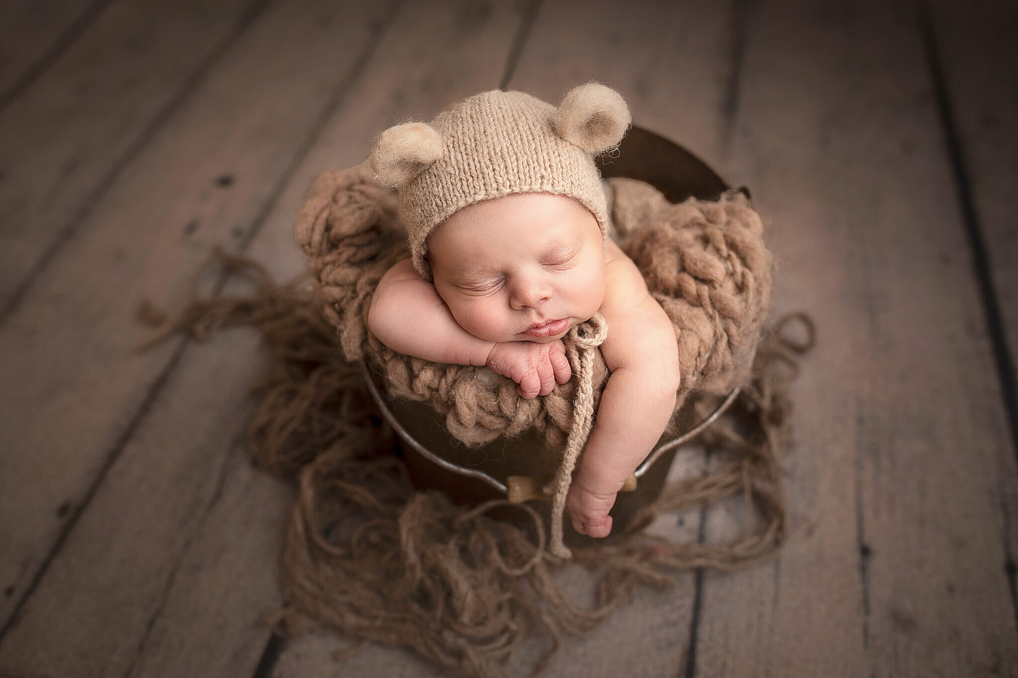 Newborn baby boy posed in bucket on grey wood floor.  Baby boy is wearing a little bear hat.