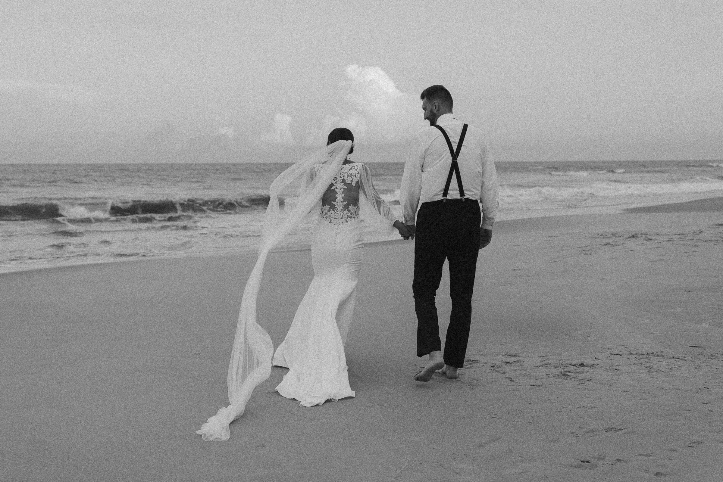Couple holding hands, walking along the beach in their wedding attire.