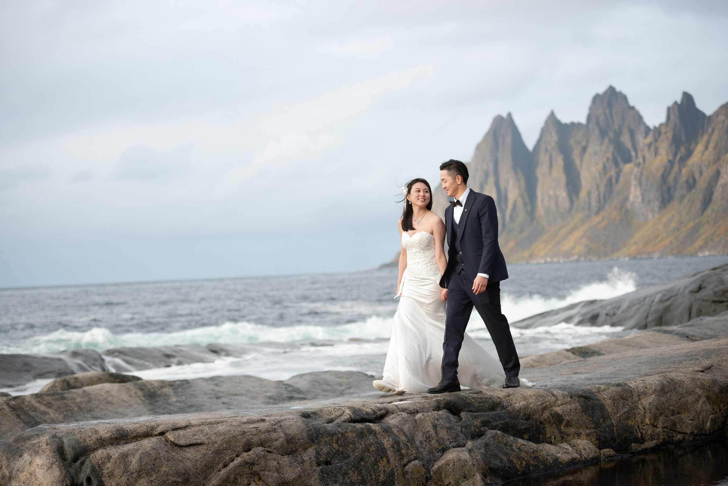 An Asian couple celebrates their elopement on the rugged shores of Senja, Norway, with the jagged teeth of the Tungeneset mountains behind them, encapsulating the wild spirit and beauty of their unique wedding day.